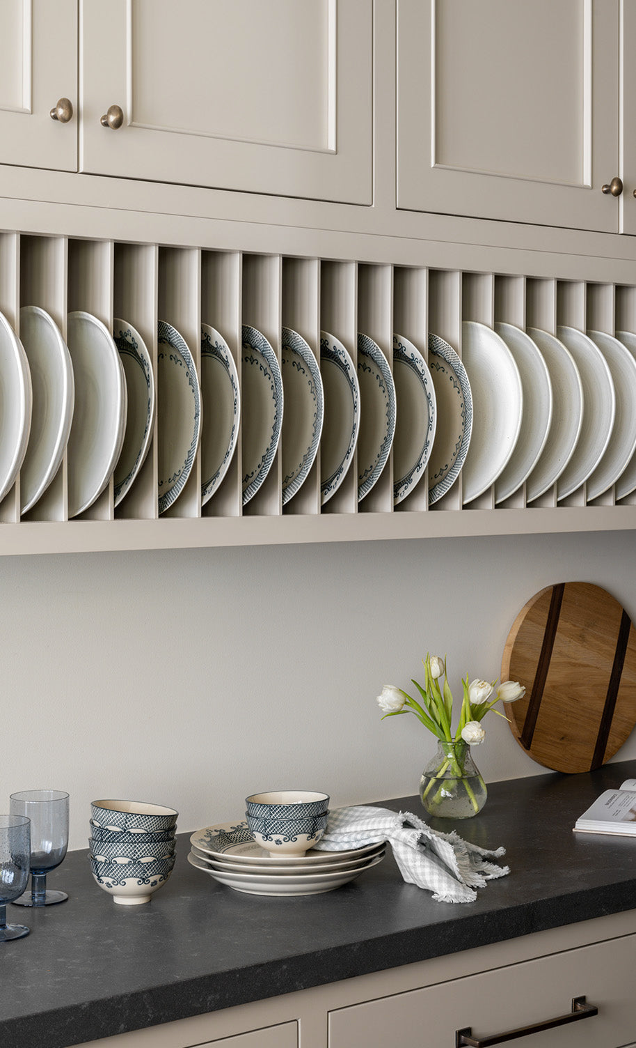 Plates are neatly arranged vertically in a rack under beige kitchen cabinets. A countertop below holds bowls, stacked plates, two blue glasses, a vase with white tulips, and a wooden cutting board, by Vendixs