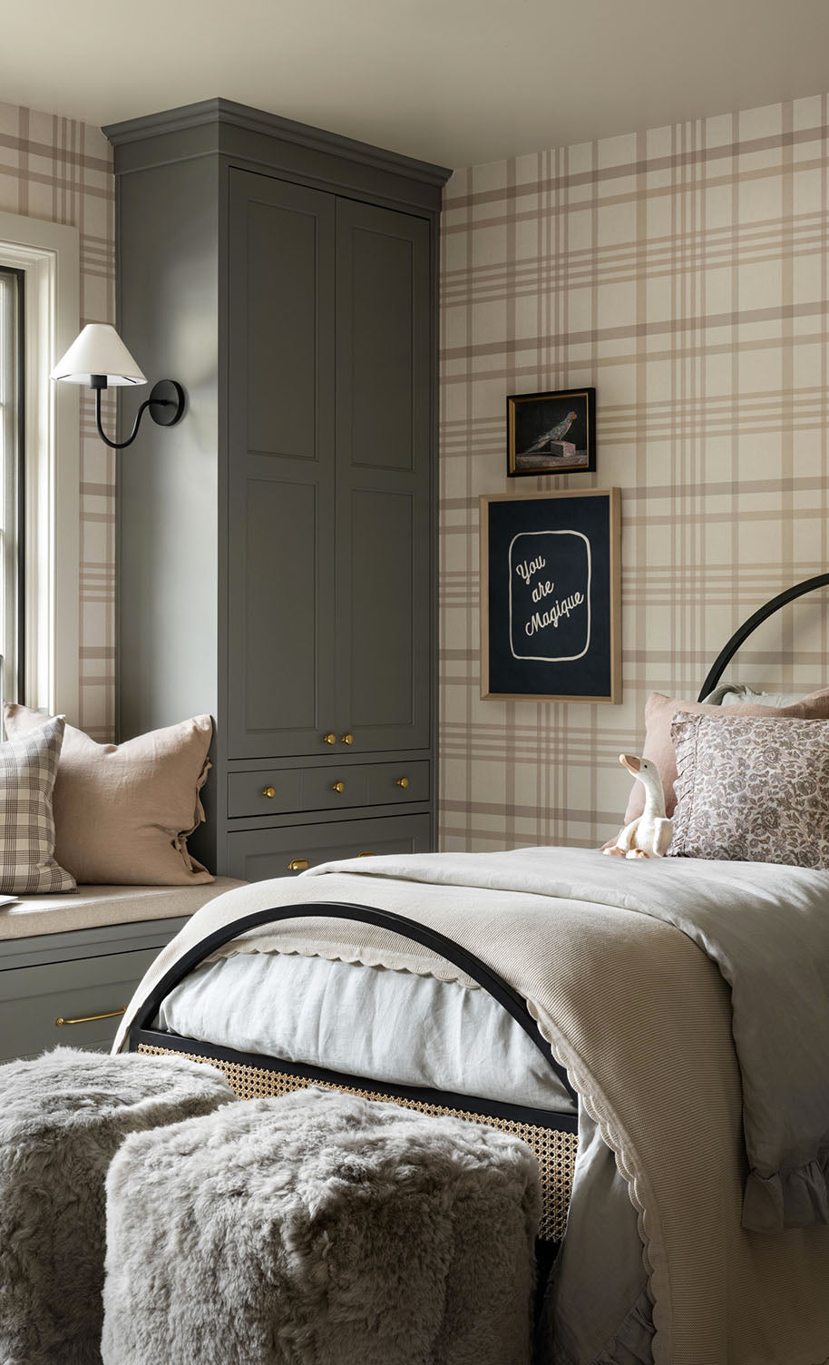 A cozy bedroom with plaid wallpaper, a bed with beige and gray bedding, a window seat with pillows, a gray wardrobe, wall art, a wall lamp, and two furry stools at the foot of the bed, by Vendixs