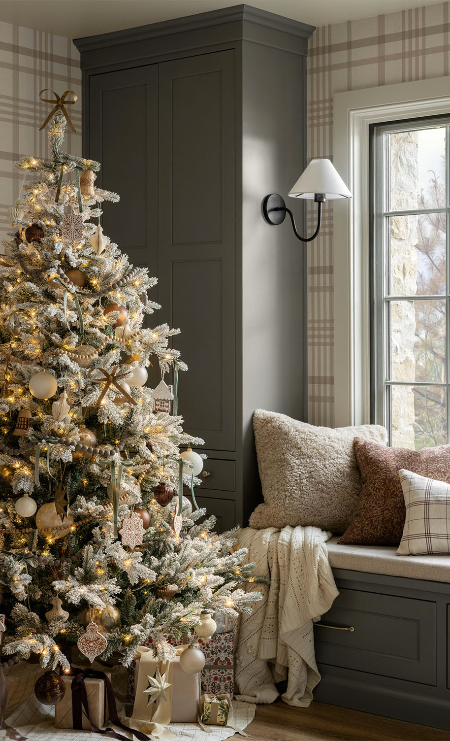 A decorated Christmas tree with lights and ornaments stands by a window seat with cozy pillows and blankets; wrapped gifts are placed under the tree in a softly lit, plaid-patterned room, by Vendixs