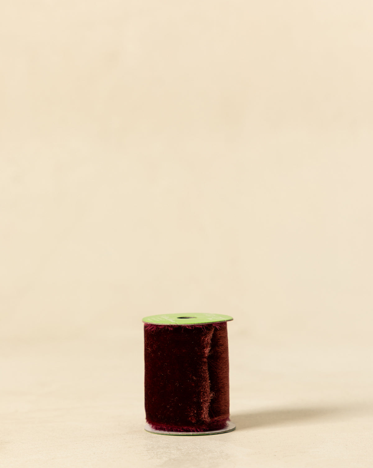 A spool of 1-3/8" Silk Velvet Ribbon with Frayed Edge in deep burgundy, topped with green and set on a light beige surface, adds elegant holiday flair to any matching backdrop, by Vendixs