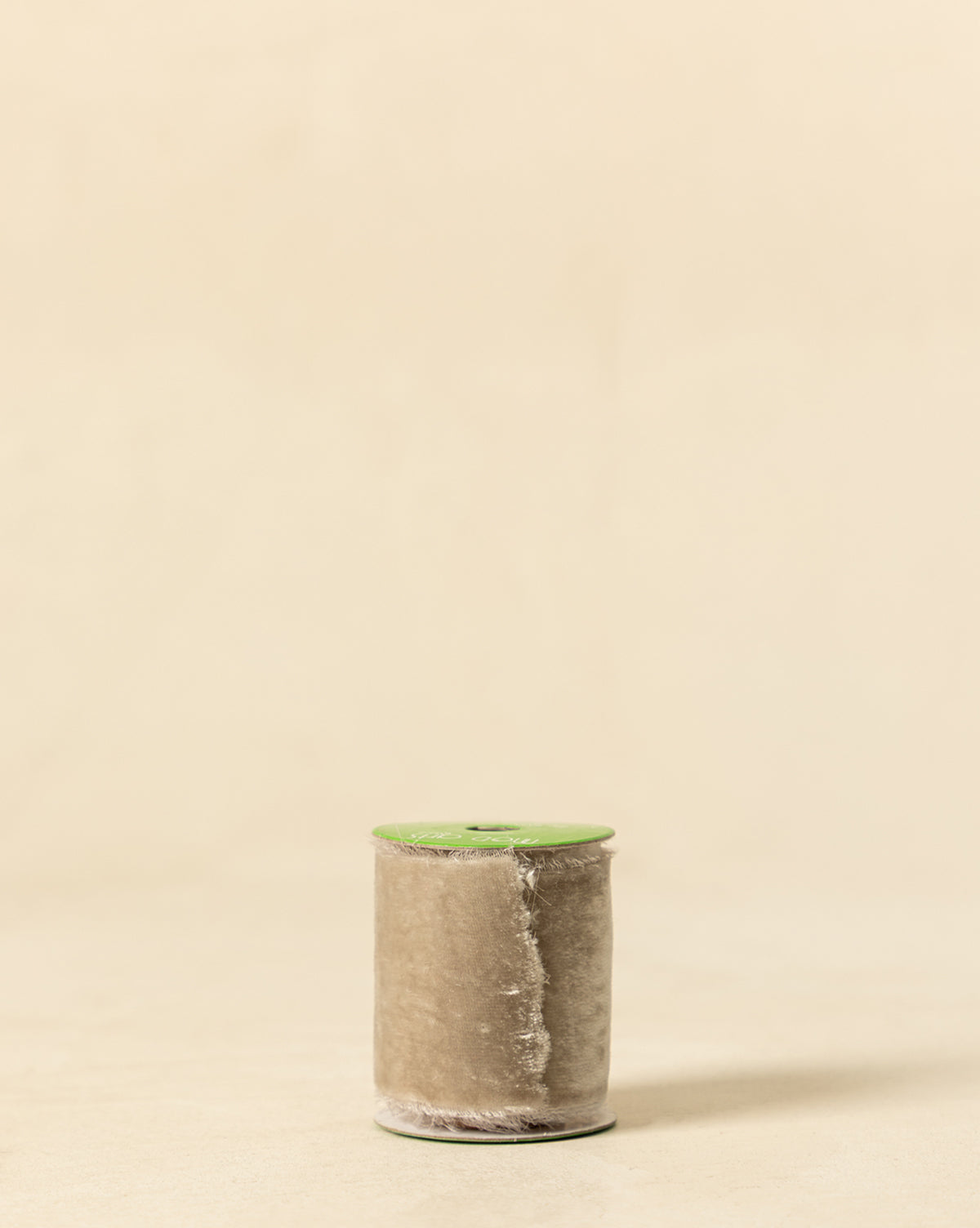 A spool of 1-3/8" Silk Velvet Ribbon with Frayed Edge in beige and a green top stands upright on a light surface, its frayed edge adding charm against the plain beige background—ideal for holiday styling, by Vendixs