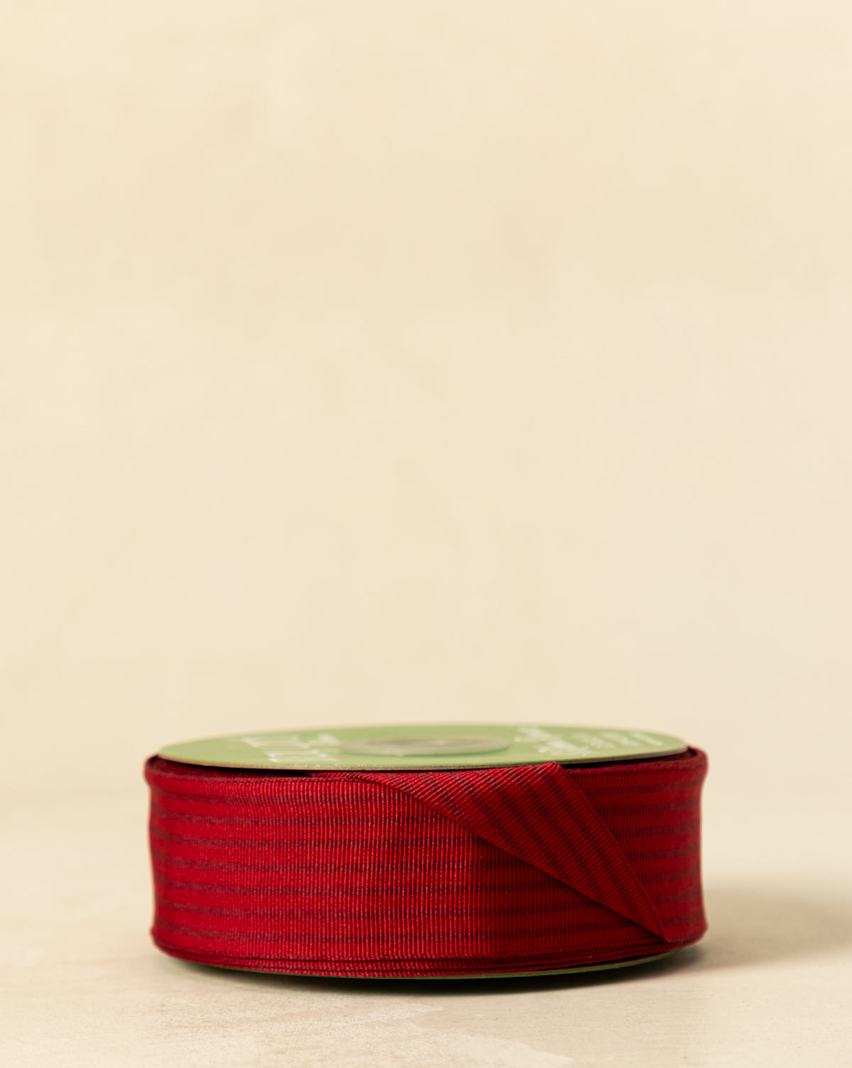 A spool of 1.5" Woven Grosgrain Stripe Ribbon with Woven Edge featuring a green top is placed on a beige surface, with some red ribbon partially unrolled and folded in front, by Vendixs