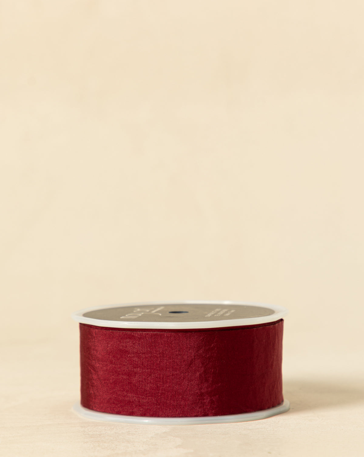 A spool of 1.5" wrinkled faux silk ribbon with cut edge, in deep red, is wrapped around a gray reel—ideal for holiday wrapping or decorating gift boxes on a light beige surface with a matching background, by McGee & Co.