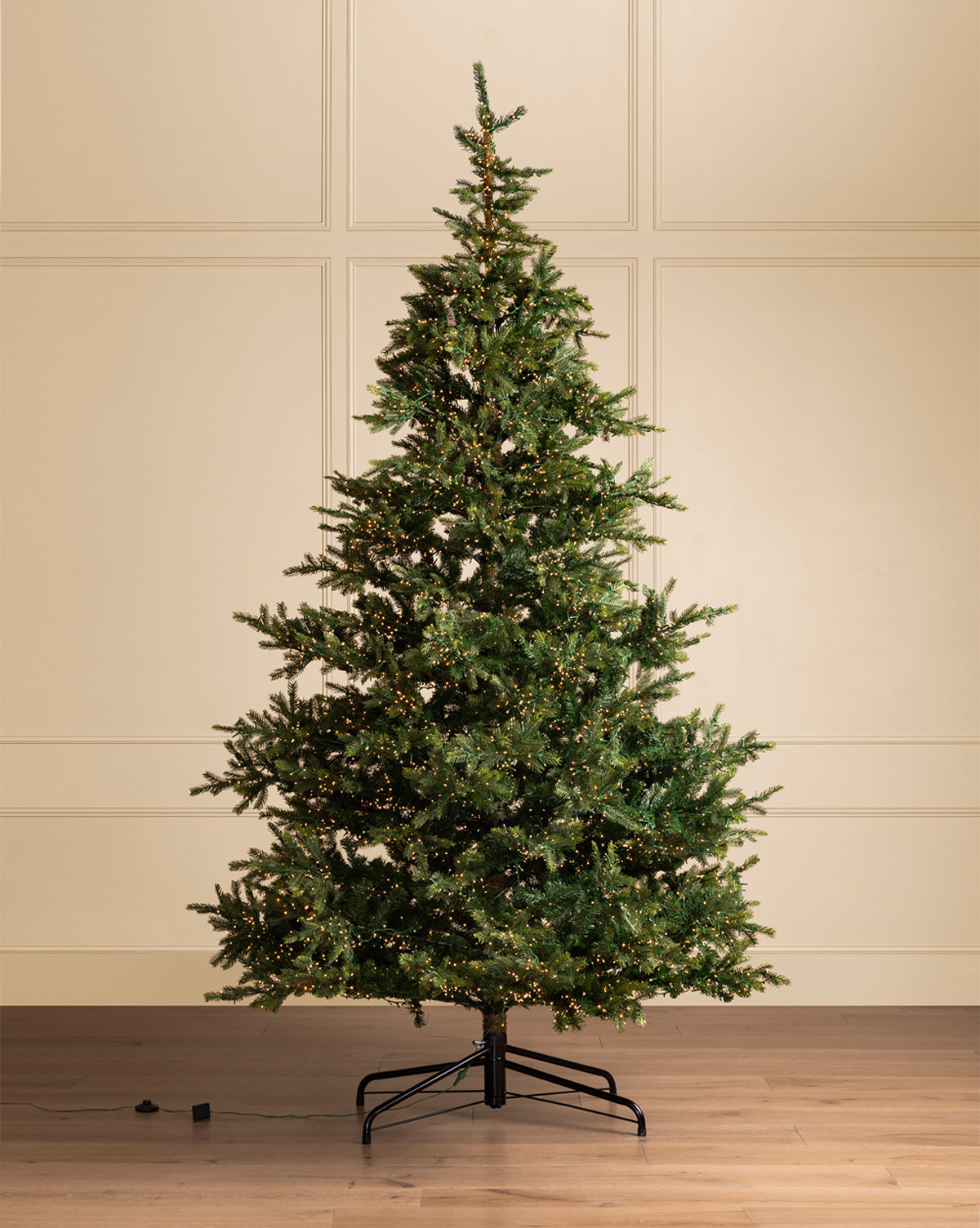A 10' Grandis Fir Pre-Lit Faux Tree stands on a wooden floor by a cream paneled wall, its LED lights twinkling across dense branches, with a power cord trailing to the left, by Vendixs
