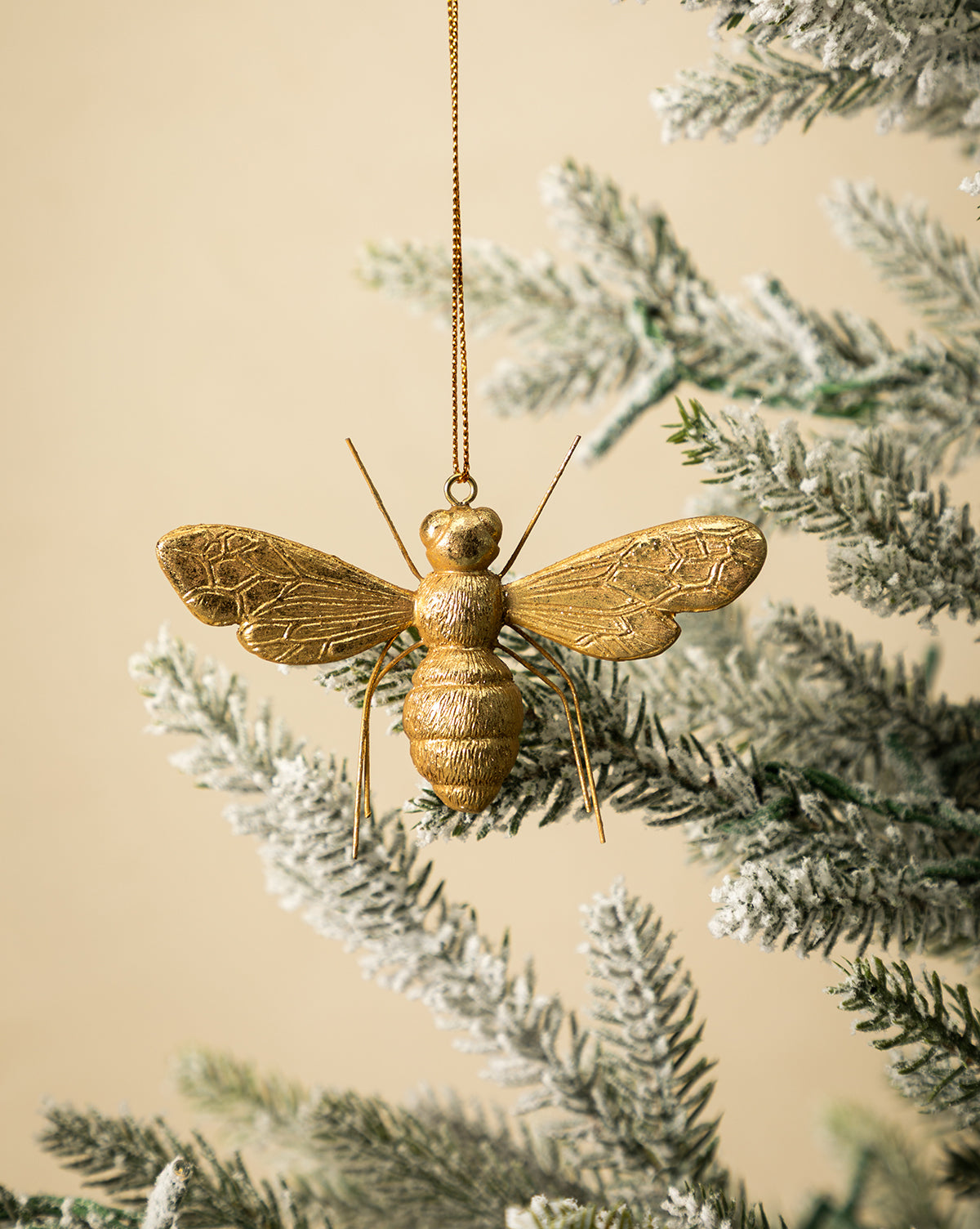 The Golden Bee Ornament, featuring a nature-inspired design, hangs from a gold string on a snow-dusted artificial holiday branch set against a light beige background, by Vendixs