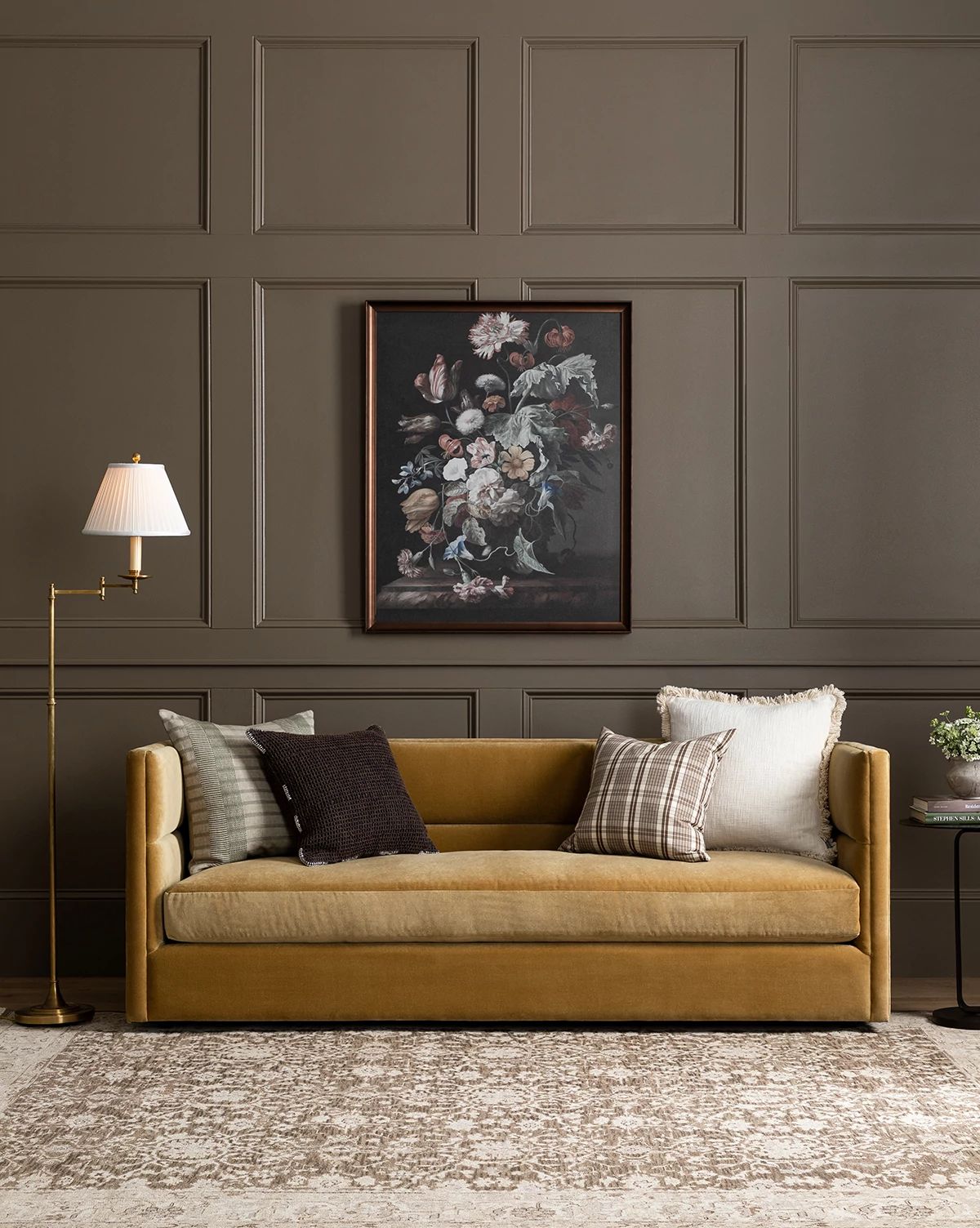 A Claremont Sofa 84 in mustard yellow, accented with assorted pillows, sits against a brown paneled wall under a floral painting. A brass floor lamp and small side table complete this cozy, elegant living room setting, by Vendixs