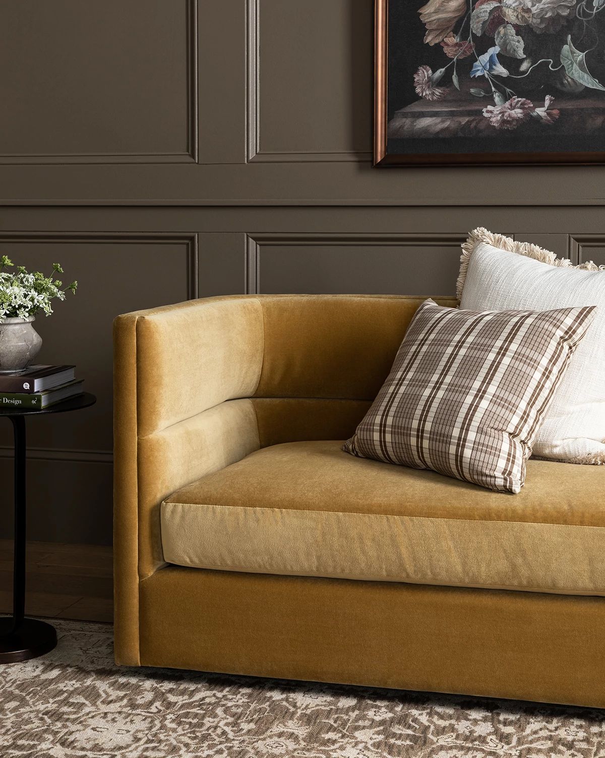 A gold velvet Claremont Sofa 84 with white and plaid pillows sits by a black side table holding a plant and books, set against dark paneled walls topped with a framed floral painting, by Vendixs