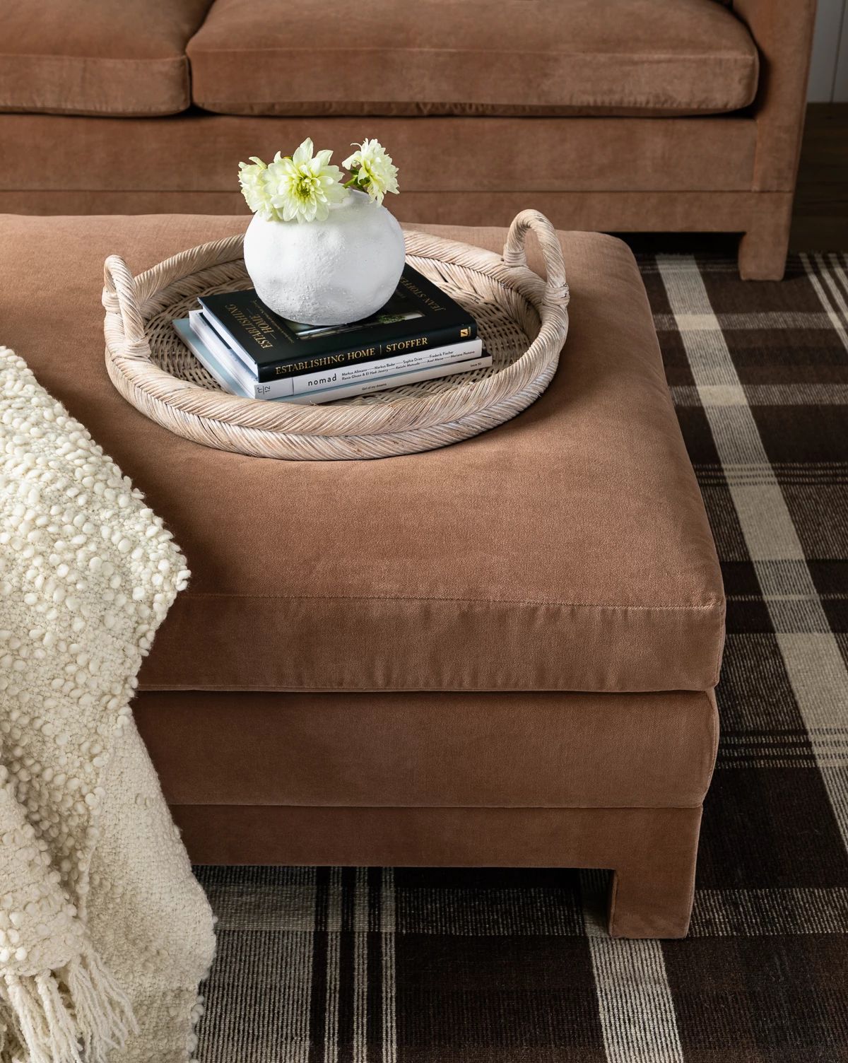 The Makerspalm Appoline Ottoman in tan is topped with a woven tray holding a white vase of flowers and two books, set next to a textured cream blanket on a brown and cream plaid rug, Vendixs