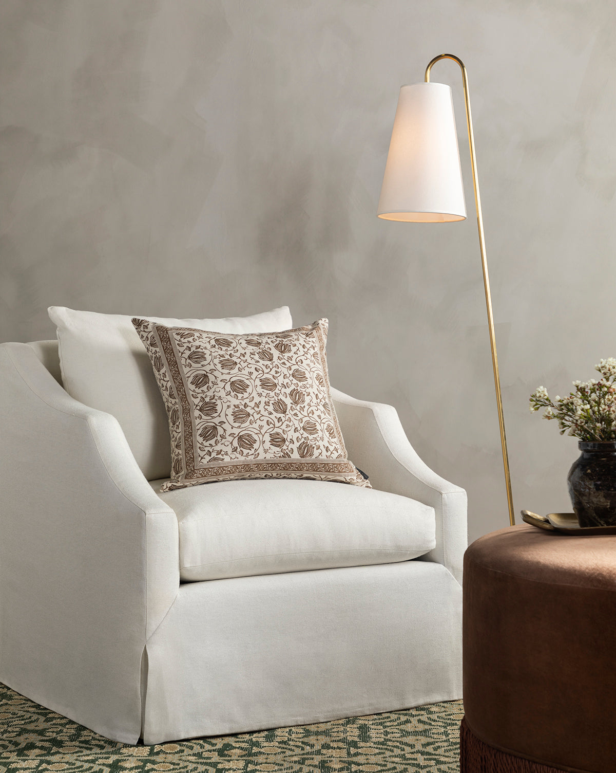 A cozy Everleigh Slipcover Lounge Chair by Community with a patterned beige cushion sits beside a gold floor lamp with a white shade, while a round brown ottoman and floral vase complete the elegant, neutral-toned decor. Vendixs