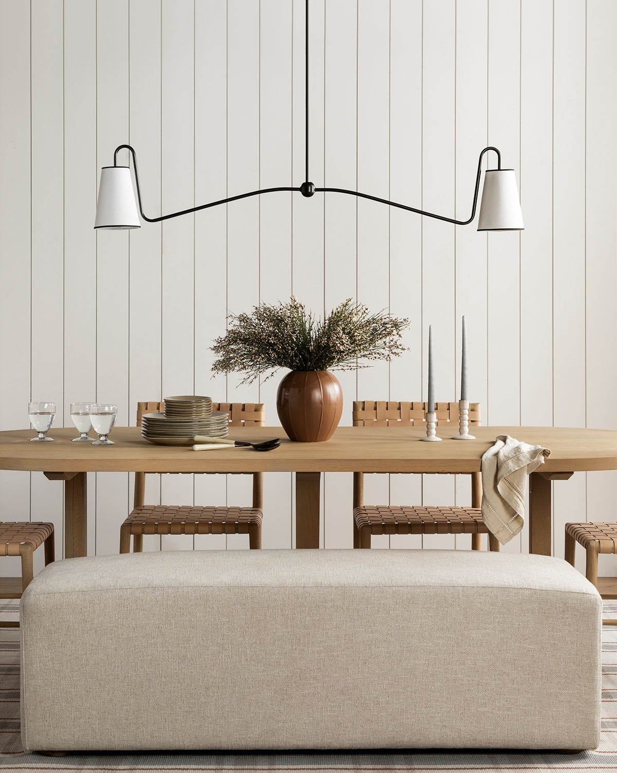 A minimalist dining room showcases a light wood table set with plates, glasses, and candles, a brown vase with greenery, woven chairs, a beige bench, and elegant lighting from the Makerspalm Jonas Linear Chandelier above. Vendixs