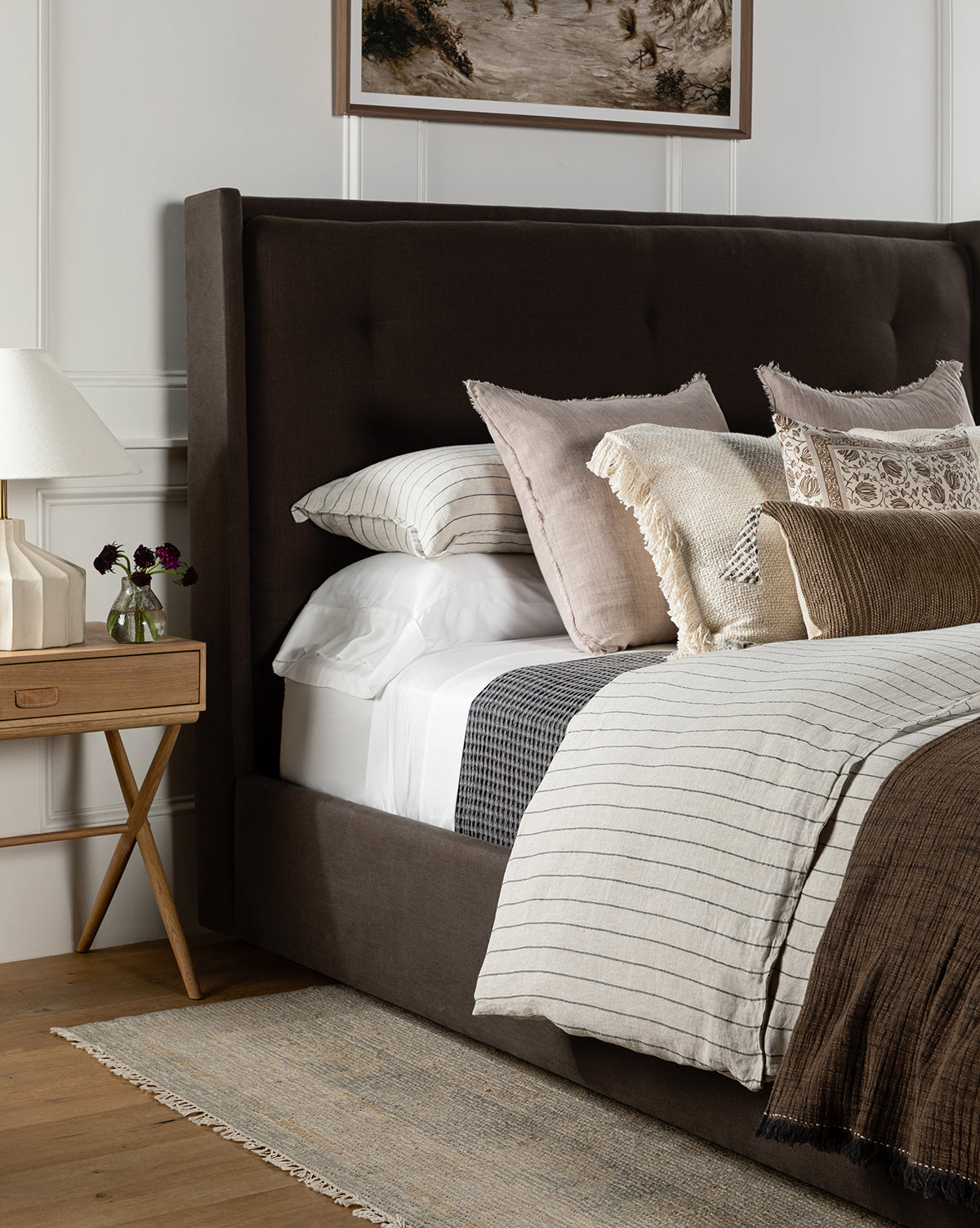 A stylish bedroom featuring the Ria Bed with a tufted headboard, layered neutral pillows and blankets, a wooden nightstand with lamp, and a small vase of flowers on a woven rug, by McGee & Co.