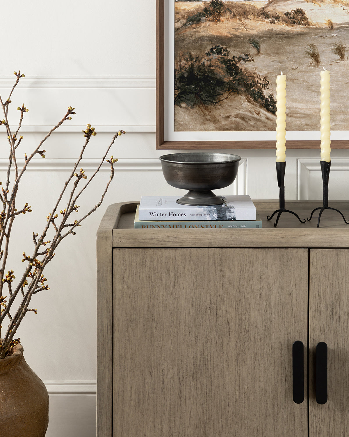 The Nyx Sideboard, made of solid mahogany with black handles, displays stacked books, a black bowl, and a candlestick with yellow candles. A potted branch is beside it, topped by a framed landscape painting above by McGee & Co.