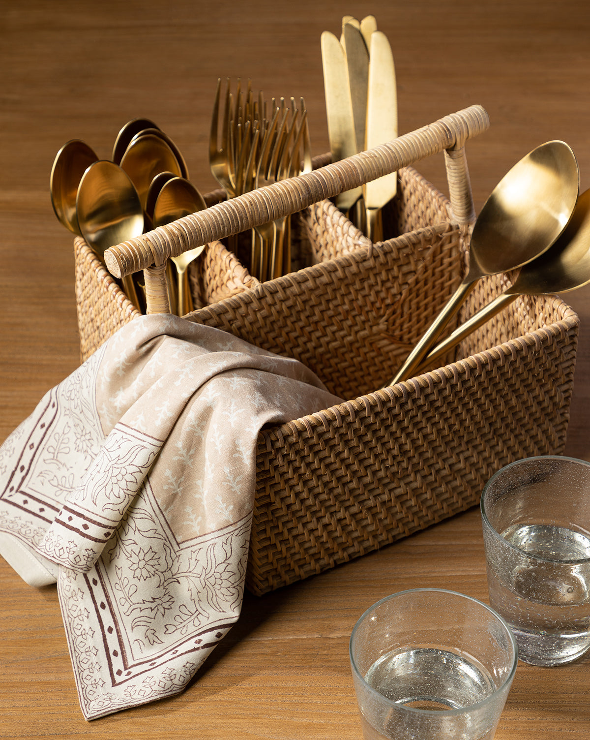 A Rattan Utensil Caddy filled with gold-colored cutlery, a patterned beige napkin over its side, and two glasses of water are arranged on a wooden table by Vendixs