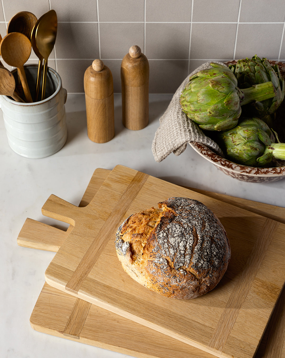 A round loaf of rustic bread sits on the Rectangle Bread Board, made from oak and walnut, stacked on natural-finish cutting boards. Nearby are artichokes, wooden salt and pepper shakers, and a ceramic utensil holder by Vendixs