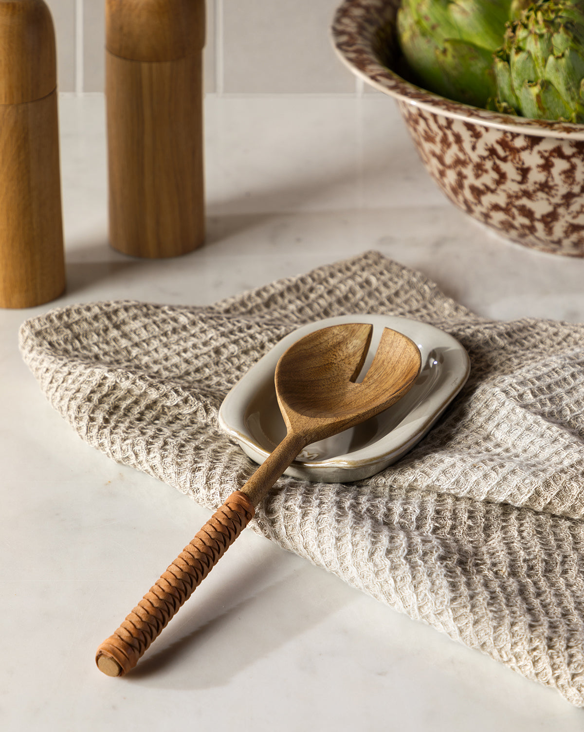 A wooden serving spoon with a woven handle rests on a small white dish and the Bunton Waffle Tea Towel. Wooden salt and pepper mills and a bowl of green artichokes sit in the background, by Vendixs