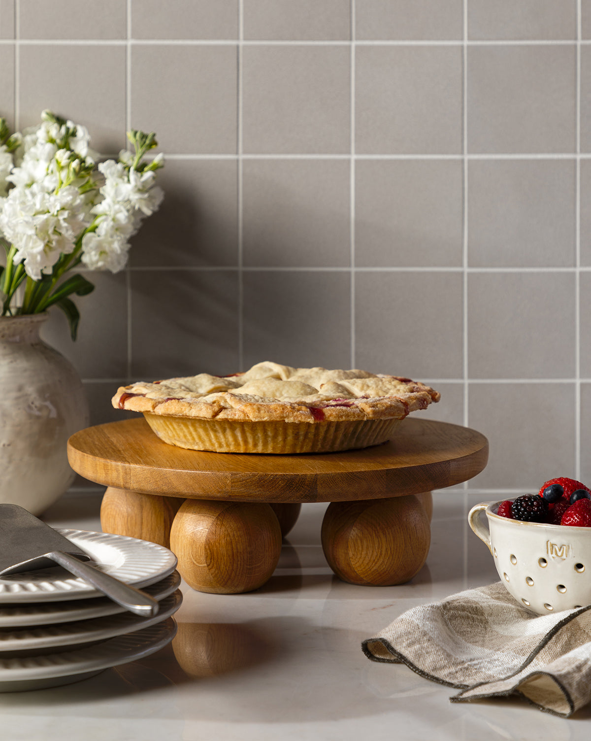 A homemade pie rests on the Bronx Oak Pedestal atop a kitchen counter, surrounded by plates, a pie server, a bowl of berries, a beige napkin, and a vase of white flowers in the background, by Vendixs