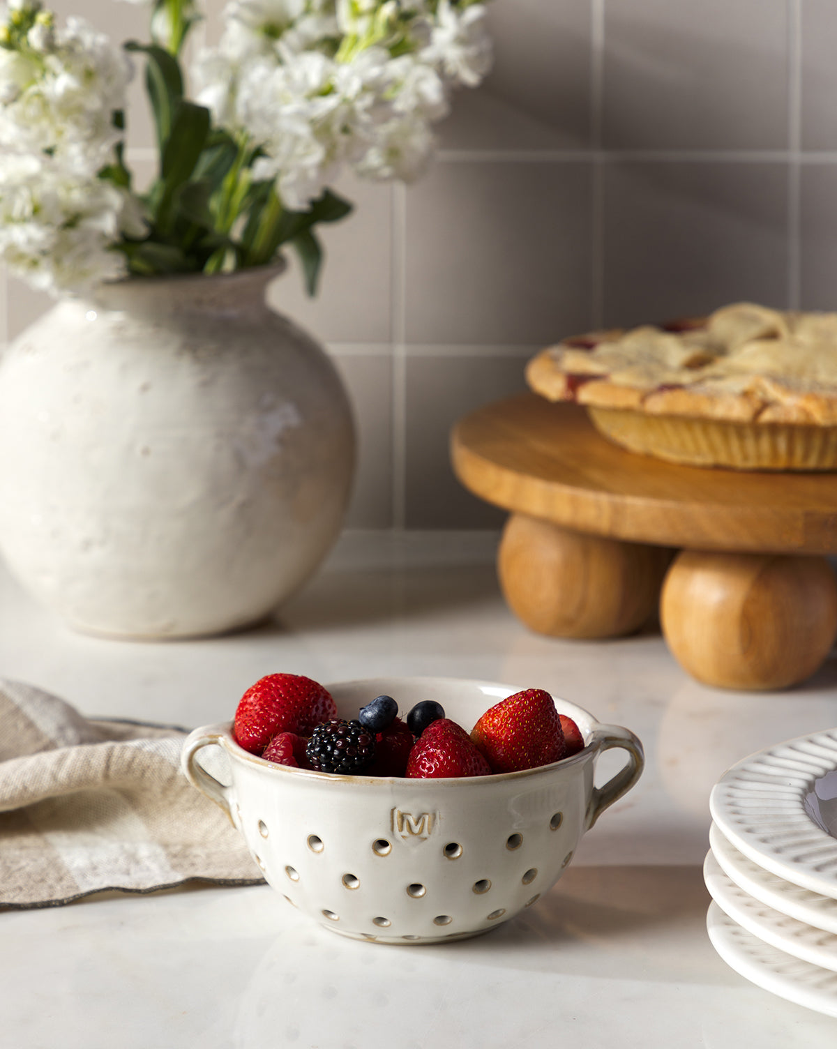 A white Glazed Berry Colander brimming with strawberries, blackberries, and blueberries sits on a kitchen counter next to stacked serveware, with a pie and white flowers enhancing the kitchen decor in the background, by Vendixs
