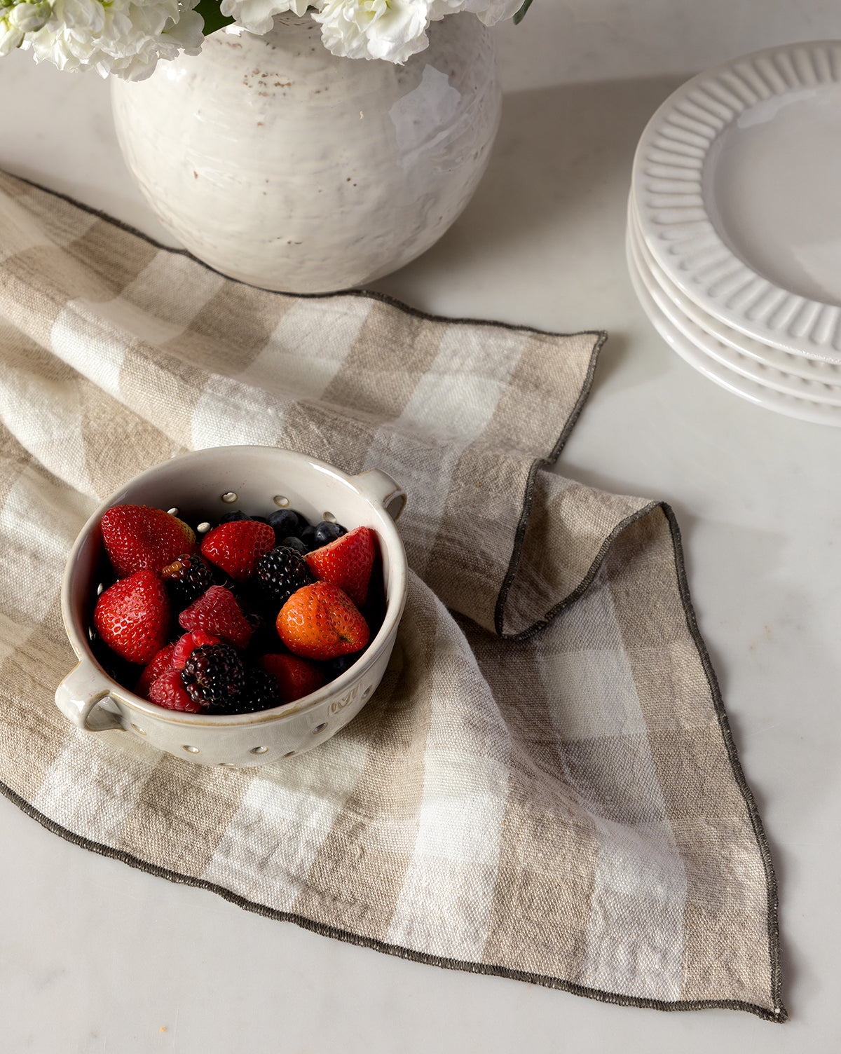 The Charlie Check Tea Towel in beige holds a white ceramic bowl of strawberries, blackberries, and blueberries beside stacked white plates and a white vase of flowers on a marble surface, by Vendixs