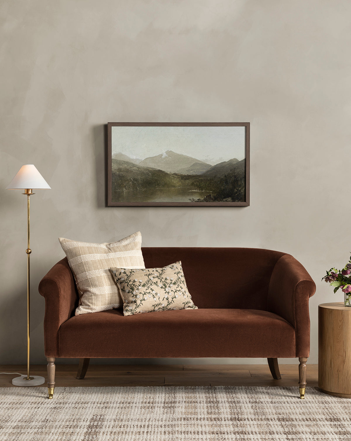 The Clemence Settee 60 (Ready to Ship), upholstered in brown and accented with patterned pillows, sits before a beige wall topped by a framed landscape. A floor lamp and side table complete this classic living room look, by Vendixs
