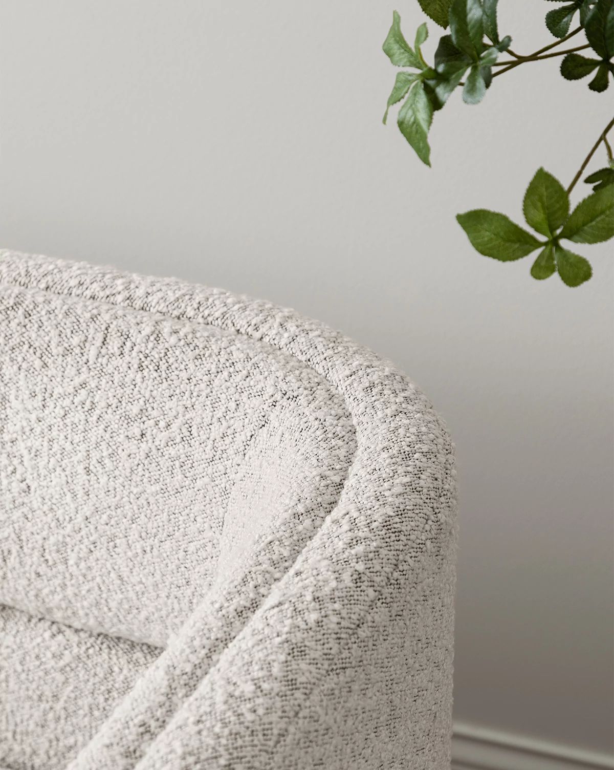 A close-up of the Claremont Modular Sofa 99 by Makerspalm shows its textured light gray upholstery and rounded edges, set against a plain wall with green plant leaves visible, highlighting its customizable modular design. Vendixs