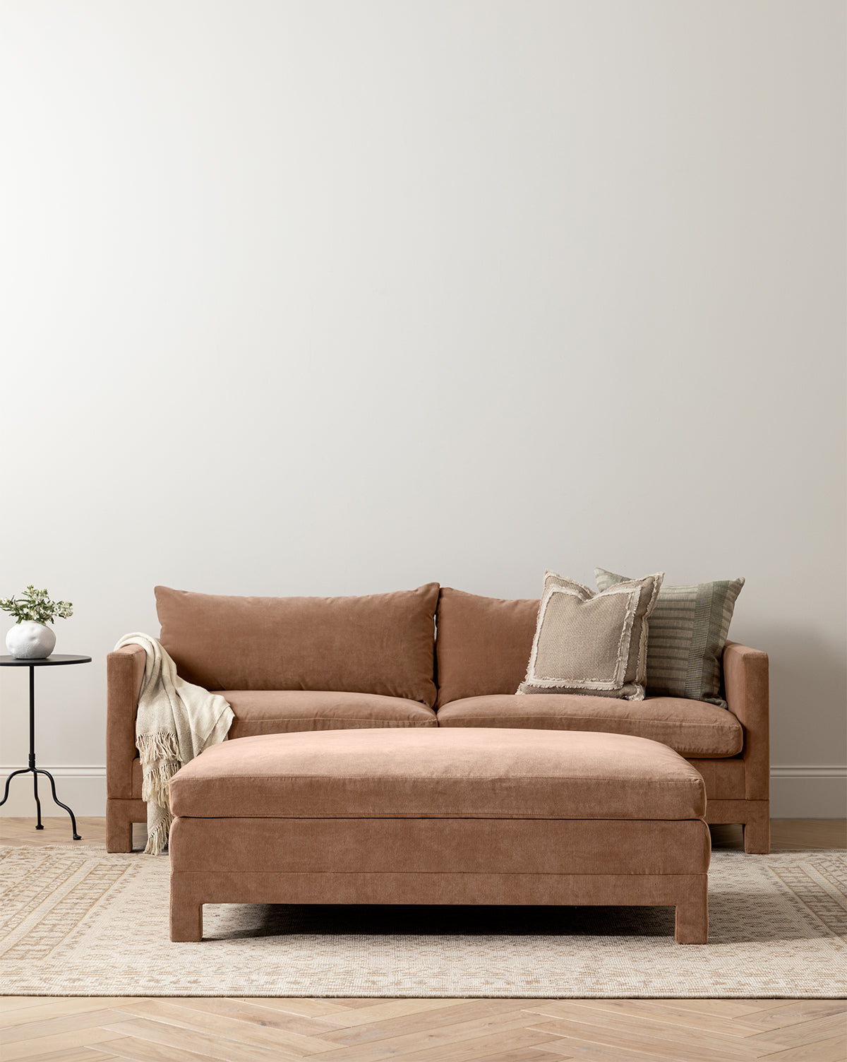 A modern living room featuring a tan sectional sofa, the made-to-order Appoline Ottoman, decorative pillows, a beige throw, a small round side table with a plant, and a light patterned rug on wooden floors, by Vendixs