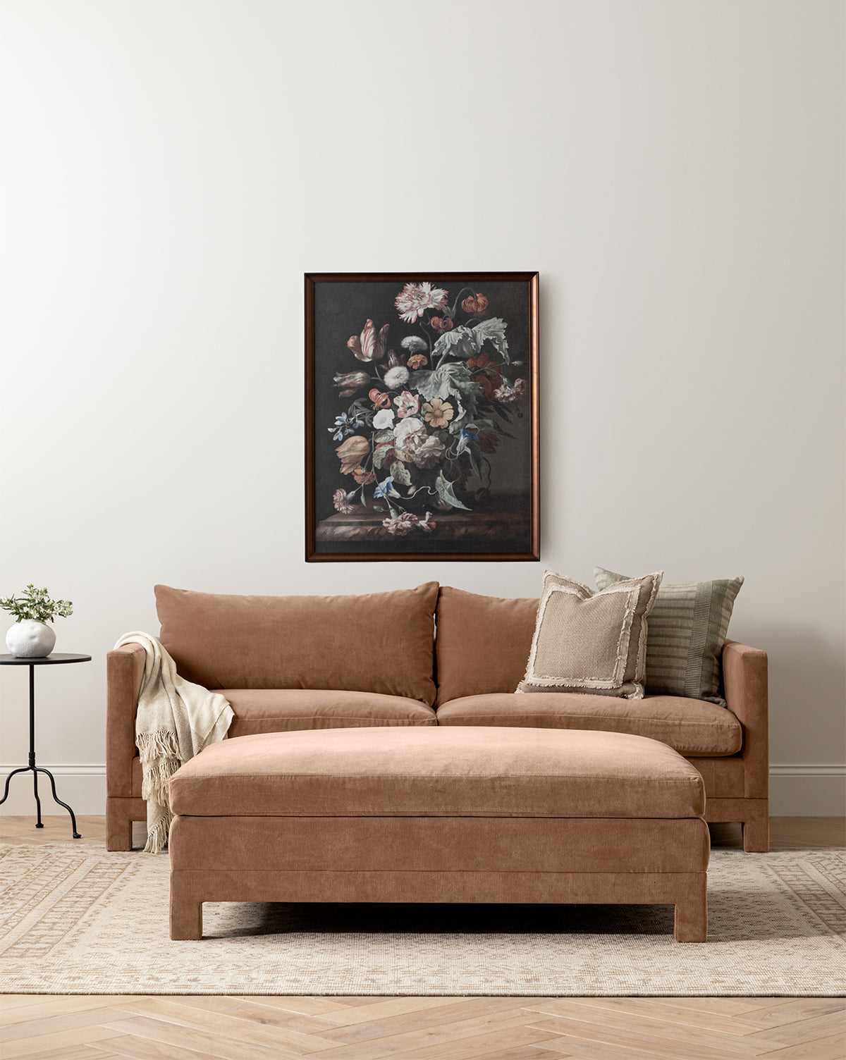 A tan Makerspalm Appoline Sofa 85 with assorted pillows and a throw anchors a minimalist living room. Above it, a floral painting hangs on the neutral wall, while a side table, plant, and beige rug complete the cozy space by Vendixs