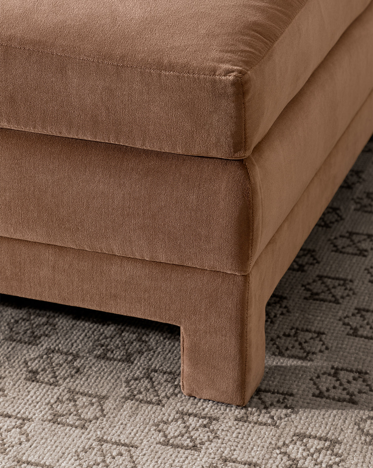 Close-up of the corner of a brown upholstered sofa with clean lines, resting on a textured gray rug next to the modern Appoline Ottoman by McGee & Co.