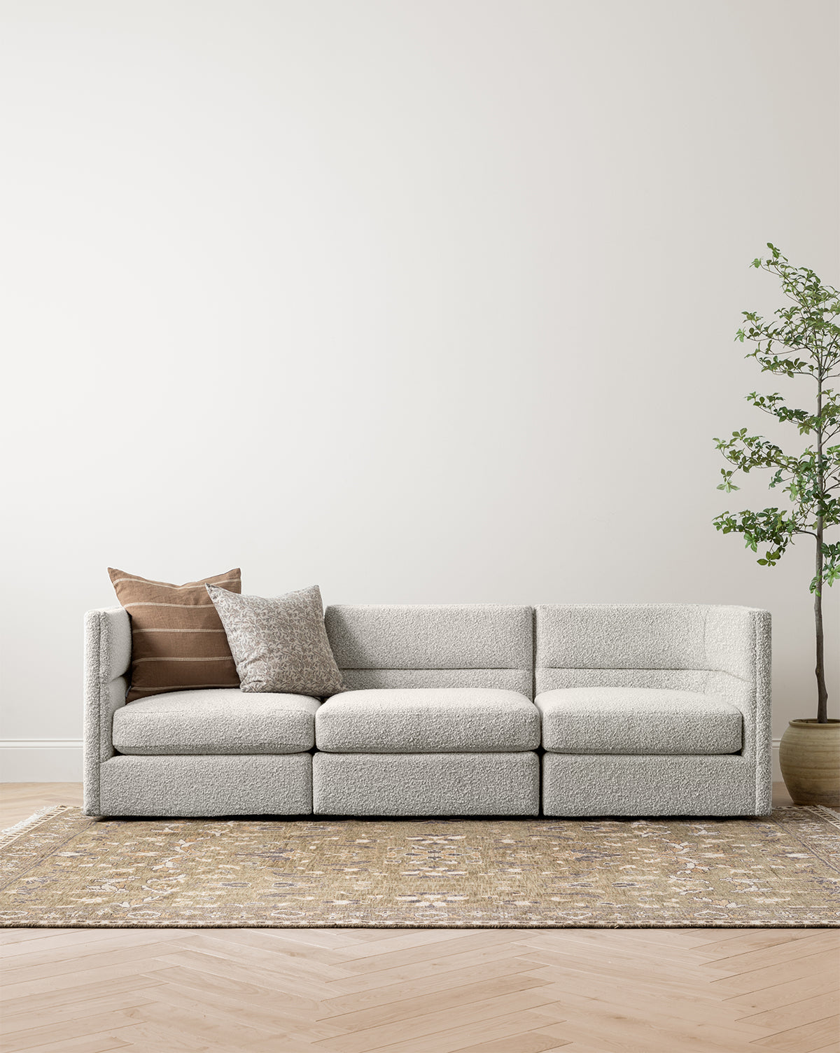 The Claremont Modular Sofa 99 by Makerspalm, featuring customizable fabric and two decorative pillows, is arranged on a patterned rug in a minimalist room with light wood flooring, cream walls, and a tall potted plant on the right, by Vendixs
