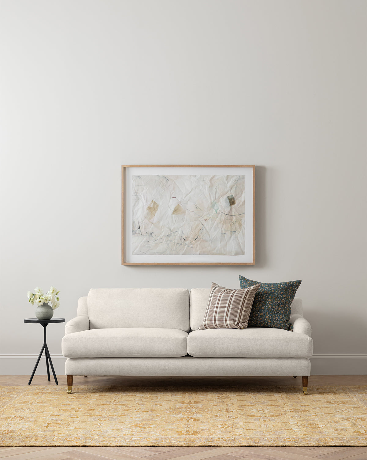 A modern living room with the customizable Lucille English Roll Arm Sofa 90.5 by Community in beige, down-wrapped cushions, two decorative pillows, a round side table with a flower vase, abstract art on a light wall, and a yellow rug on wood floors by Vendixs