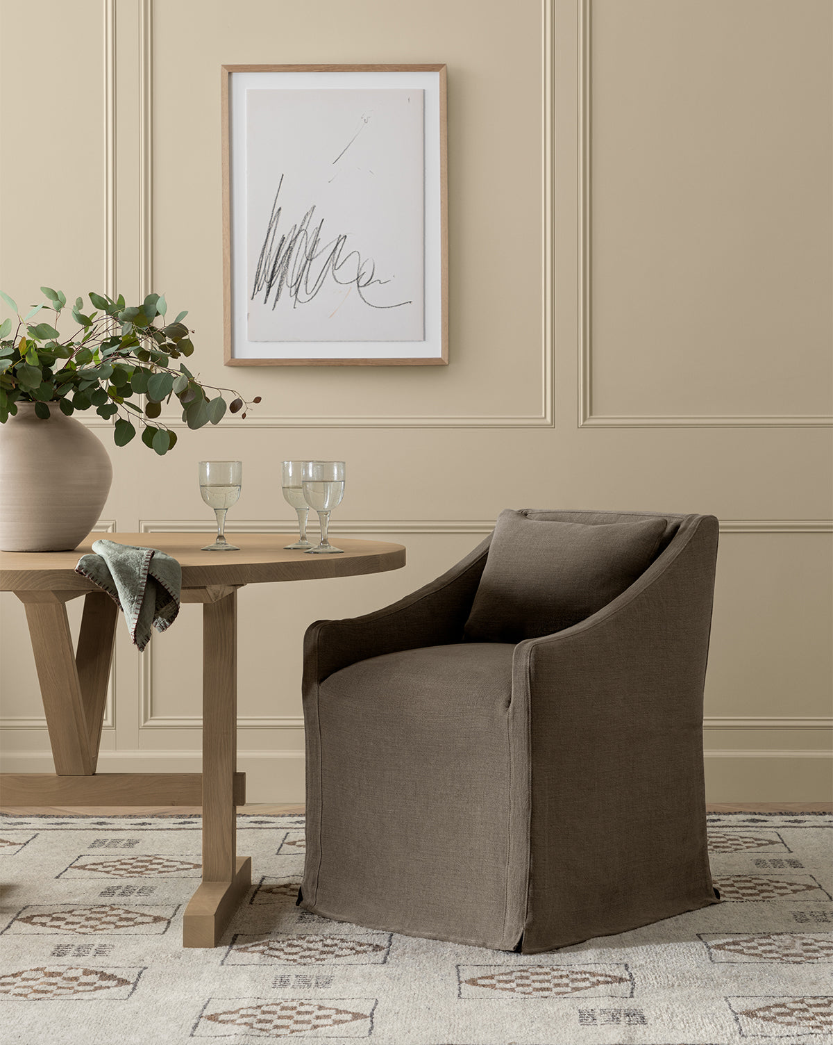 An Olivier Slipcover Dining Arm Chair in brown stands by a round wooden table set with two glasses, greenery in a vase, and a napkin. Abstract art hangs on the beige paneled wall above a patterned rug, by McGee & Co.