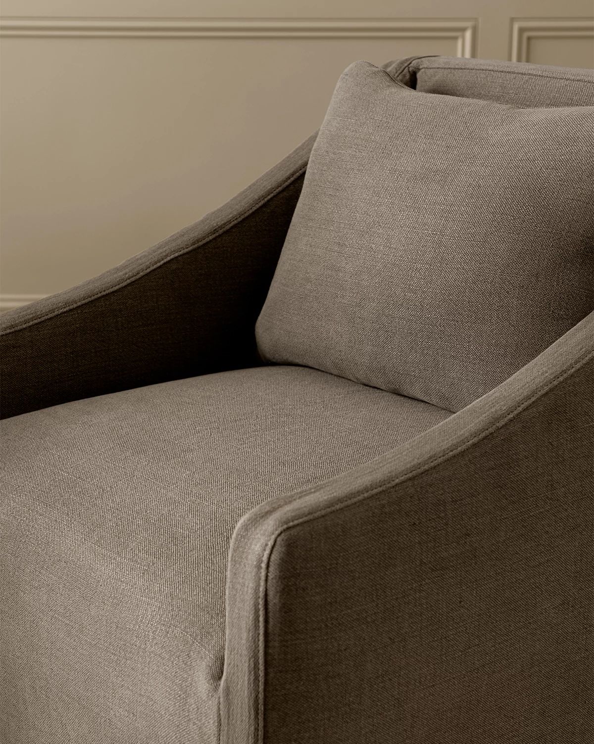 A close-up of the Olivier Slipcover Dining Arm Chair, featuring soft, textured taupe upholstery and a feather and down cushion, set against a beige paneled wall, by McGee & Co.
