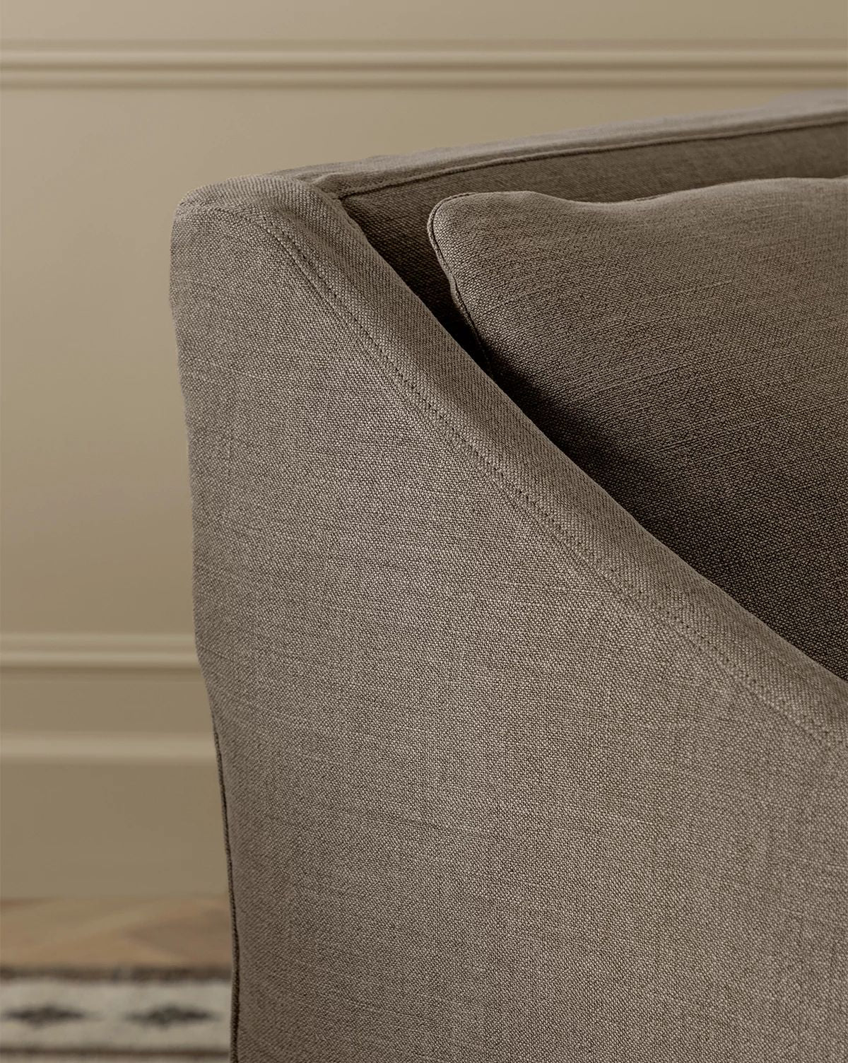 Close-up of the arm and backrest of the Olivier Slipcover Dining Arm Chair, upholstered in taupe fabric with a feather and down cushion, set against a beige wall with decorative molding. A patterned rug appears at the bottom left, by McGee & Co.