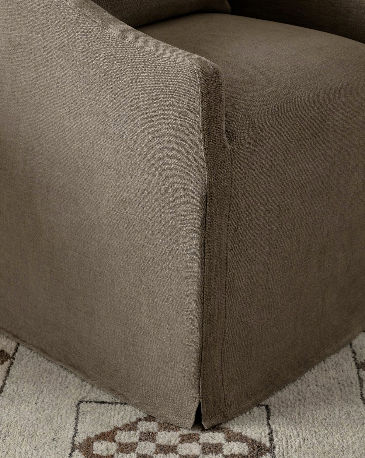 Close-up of the arm and base of the Olivier Slipcover Dining Arm Chair, upholstered in brown. The chair is positioned on a light-colored rug featuring geometric patterns in beige and brown, by McGee & Co.