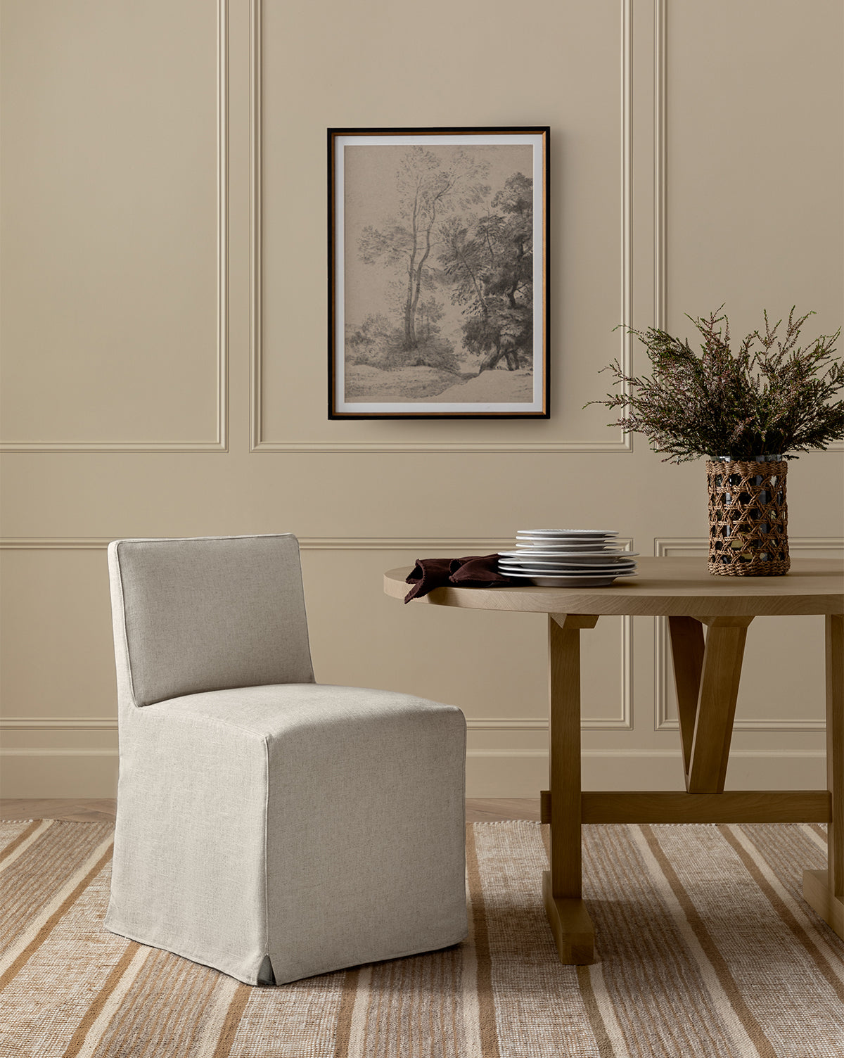 A minimalist room features the Olivier Slipcover Dining Chair, a wooden table with stacked plates and a vase of greenery, a striped rug, paneled walls, and a framed landscape artwork above, by Vendixs