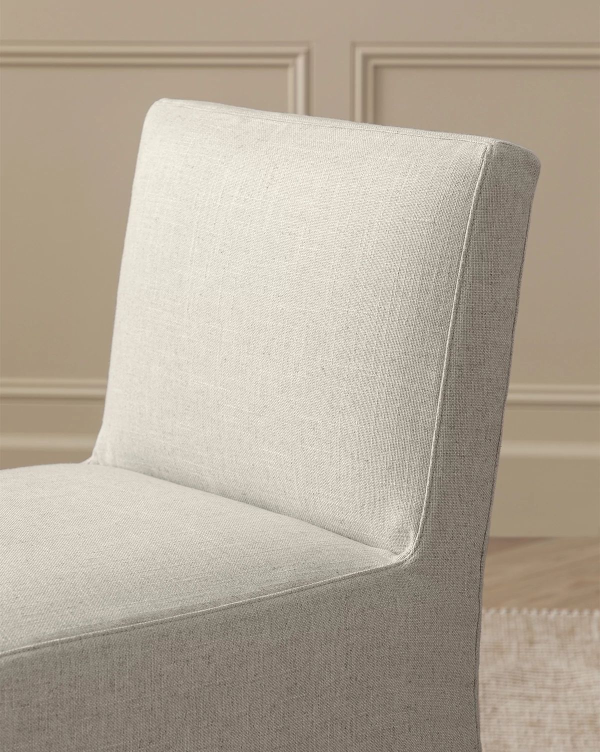 A close-up of the Olivier Slipcover Dining Chair in light beige fabric, with a feather and down cushion, set against a beige wall with decorative molding and part of a woven rug visible on the floor, by Vendixs