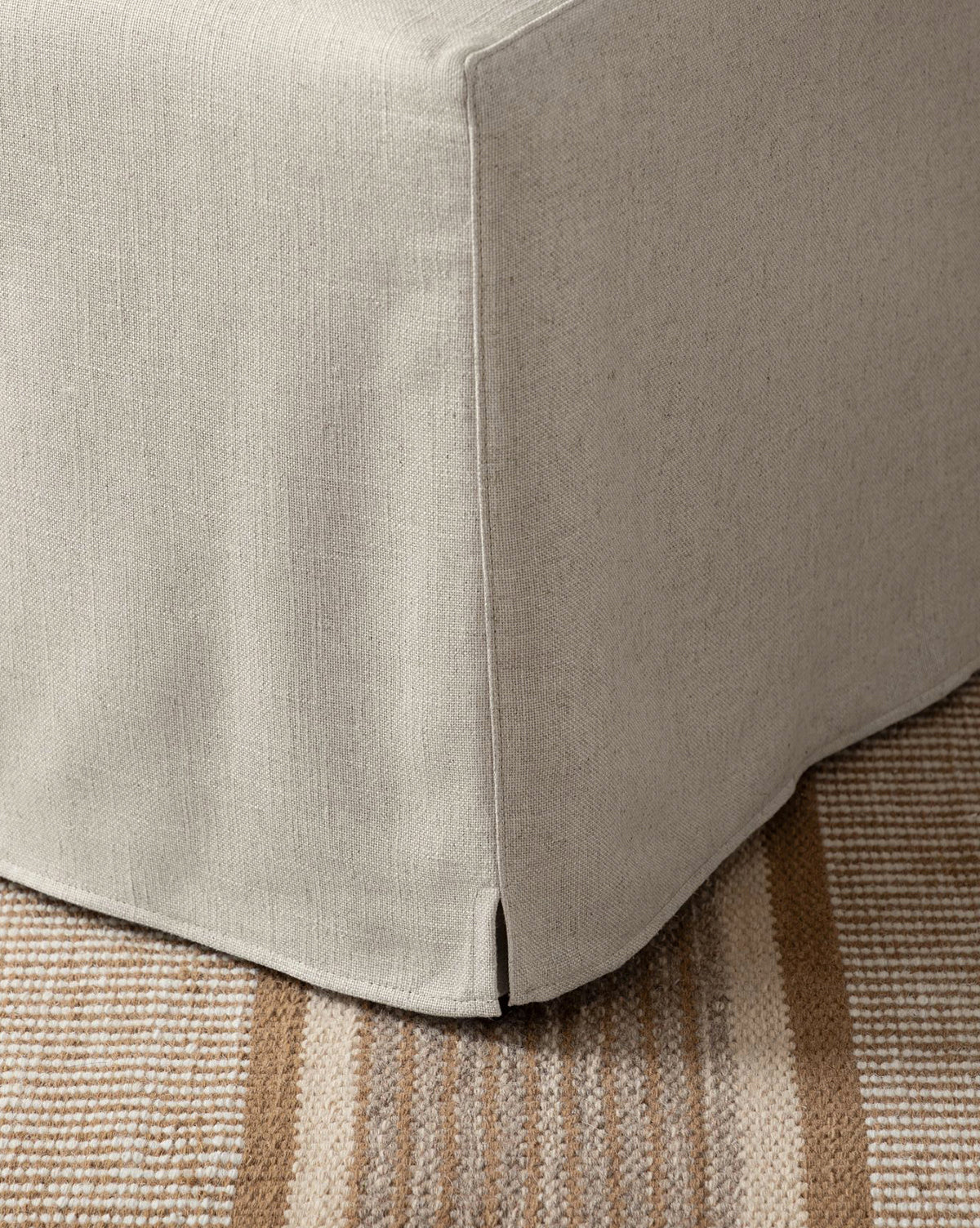 Close-up of the bottom corner of the Olivier Slipcover Dining Chair in light beige, featuring a small hem slit. The chair rests on a textured tan and brown striped rug, highlighting its stylish upholstery by McGee & Co.