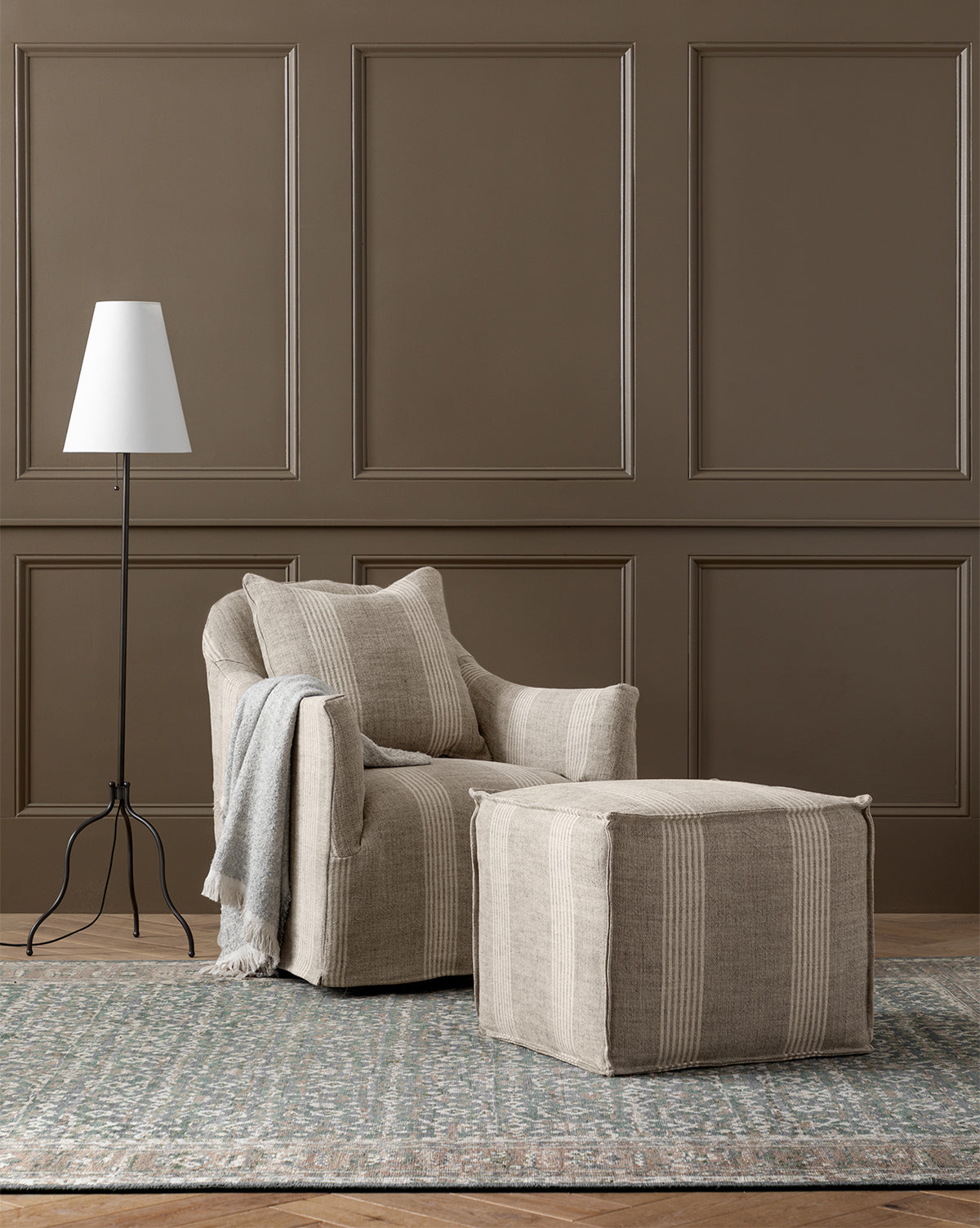 A cozy beige armchair from the made-to-order Johannes Cube collection, with a cushioned seat and matching ottoman, sits on a patterned rug by a modern white floor lamp against a brown paneled wall. A folded gray throw rests on the chair’s arm, by Vendixs
