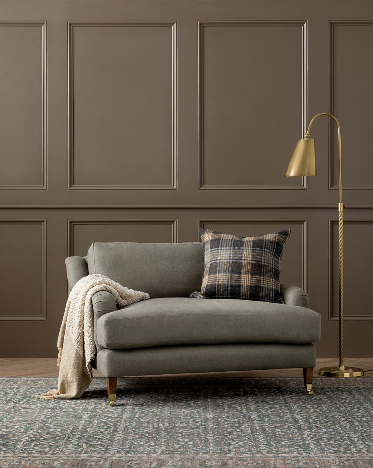 The Community Lucille English Roll Arm Chair and a Half features plush down-wrapped cushions. Shown in cozy gray with a plaid pillow and cream knit blanket, its set on a patterned rug beside a gold floor lamp and brown paneled wall. Vendixs