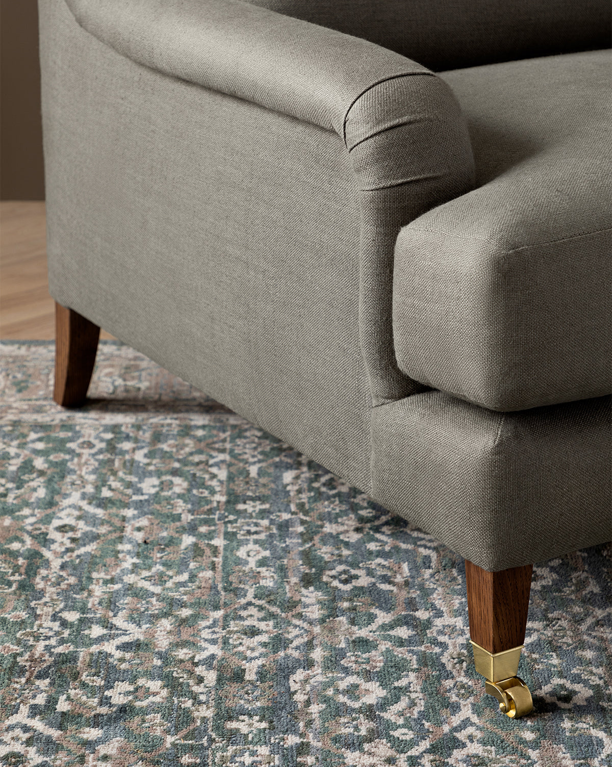 Close-up of a gray upholstered sofa with wooden legs and brass casters, beside the Lucille English Roll Arm Chair and a Half with a plush down-wrapped cushion, all set on a blue, beige, and green patterned area rug, by Vendixs