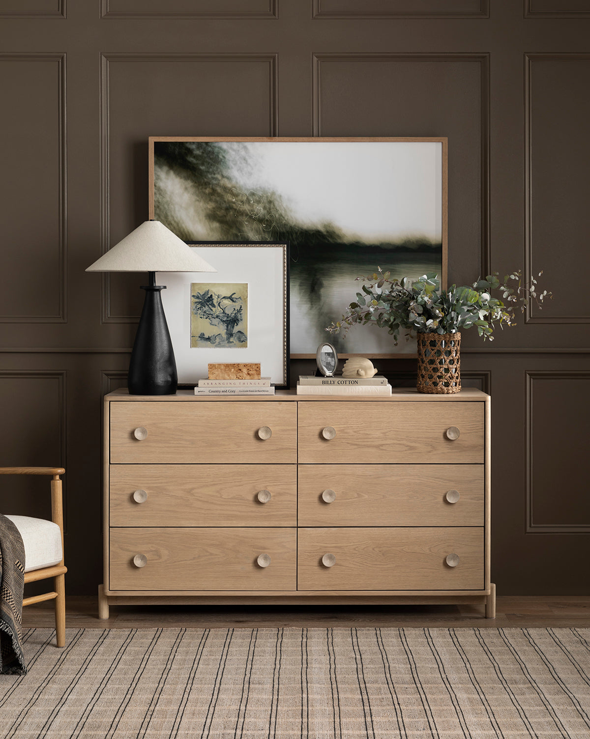 A light wood dresser with six drawers stands against a brown paneled wall beneath a rectangle mirror. On top are the Innes Tapered Table Lamp, framed art, books, a round clock, and greenery. A neutral rug and part of a wooden chair are visible by Vendixs