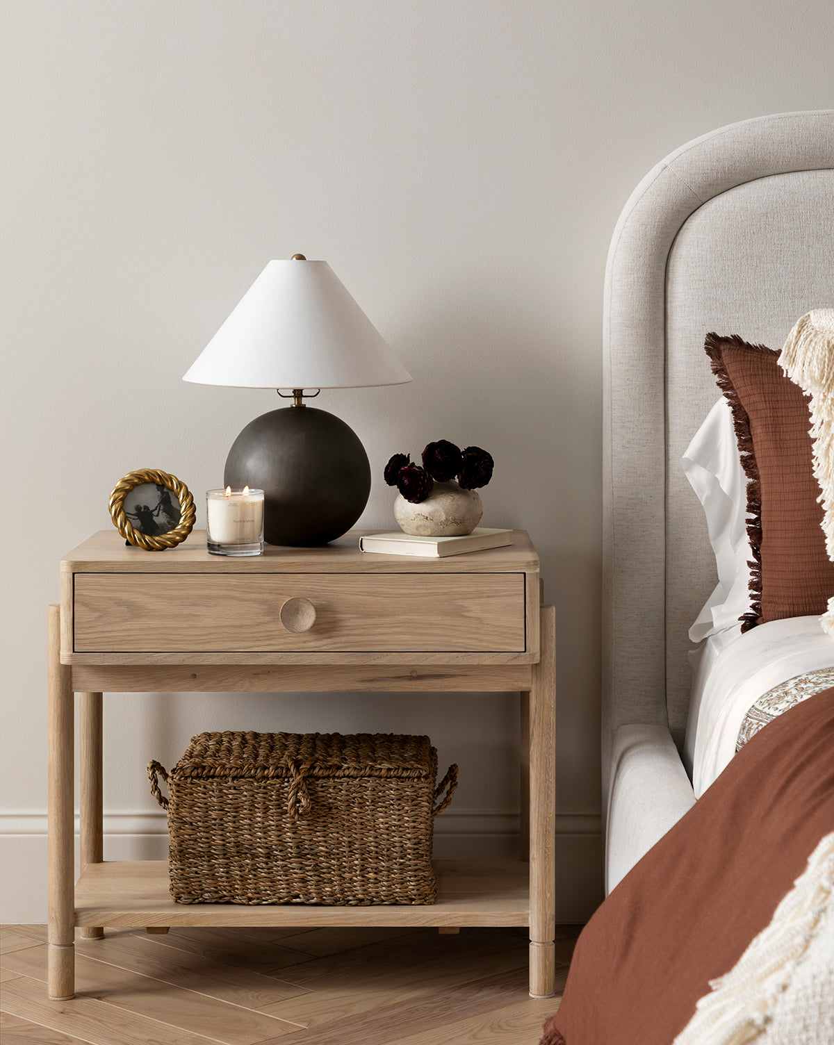 A wooden nightstand beside a bed holds a black lamp, candle, photo frame, and vase with dark flowers. The Kya Seagrass Box sits on the lower shelf. The bedding is brown and cream, by Vendixs
