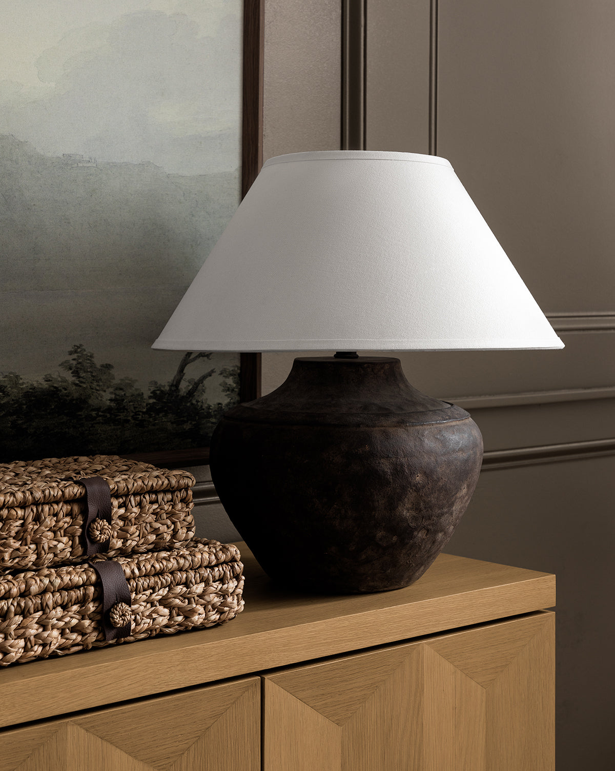 The Gannon Table Lamp, crafted from dark ceramic with a fabric shade, sits on a wooden sideboard beside stacked woven baskets, framed by a landscape painting and paneled wall in the background, by McGee & Co.