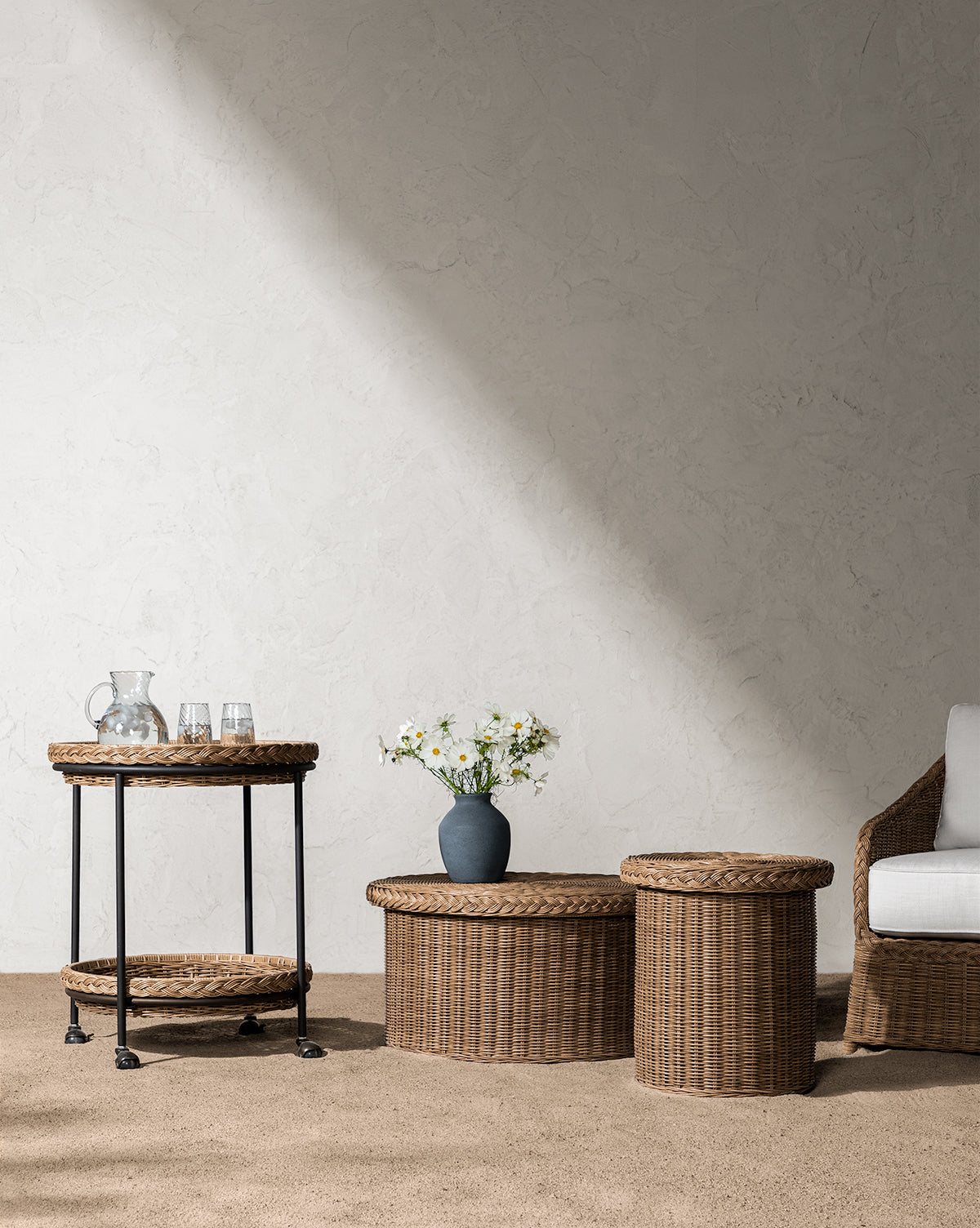 A minimalist room showcases the weather-resistant Haviland Side Table, a bar cart with a glass pitcher and glasses, two wicker tables, a vase of white flowers, and a white cushioned chair against a textured wall lit by sunlight, by Vendixs