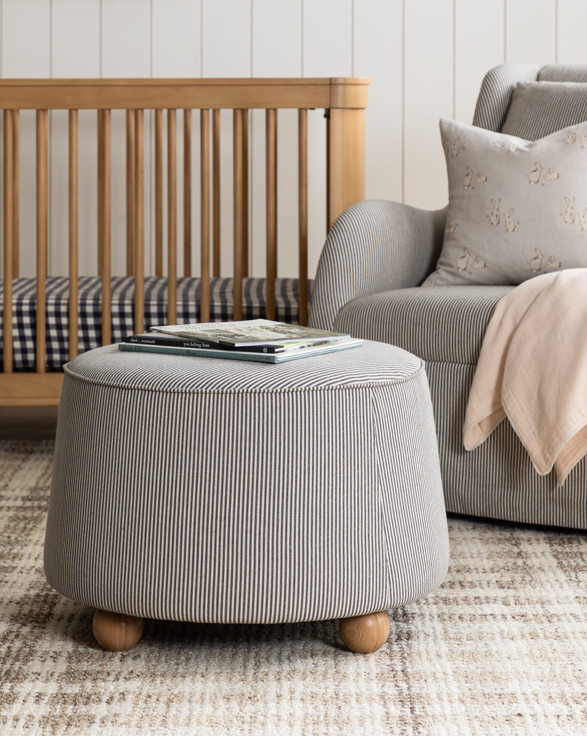 A cozy living room featuring the Barnaby Ottoman with stripes, a gray armchair and pink blanket, a wooden crib, and a patterned rug. Books sit on the ottoman amid decor in neutral and soft tones, by Vendixs