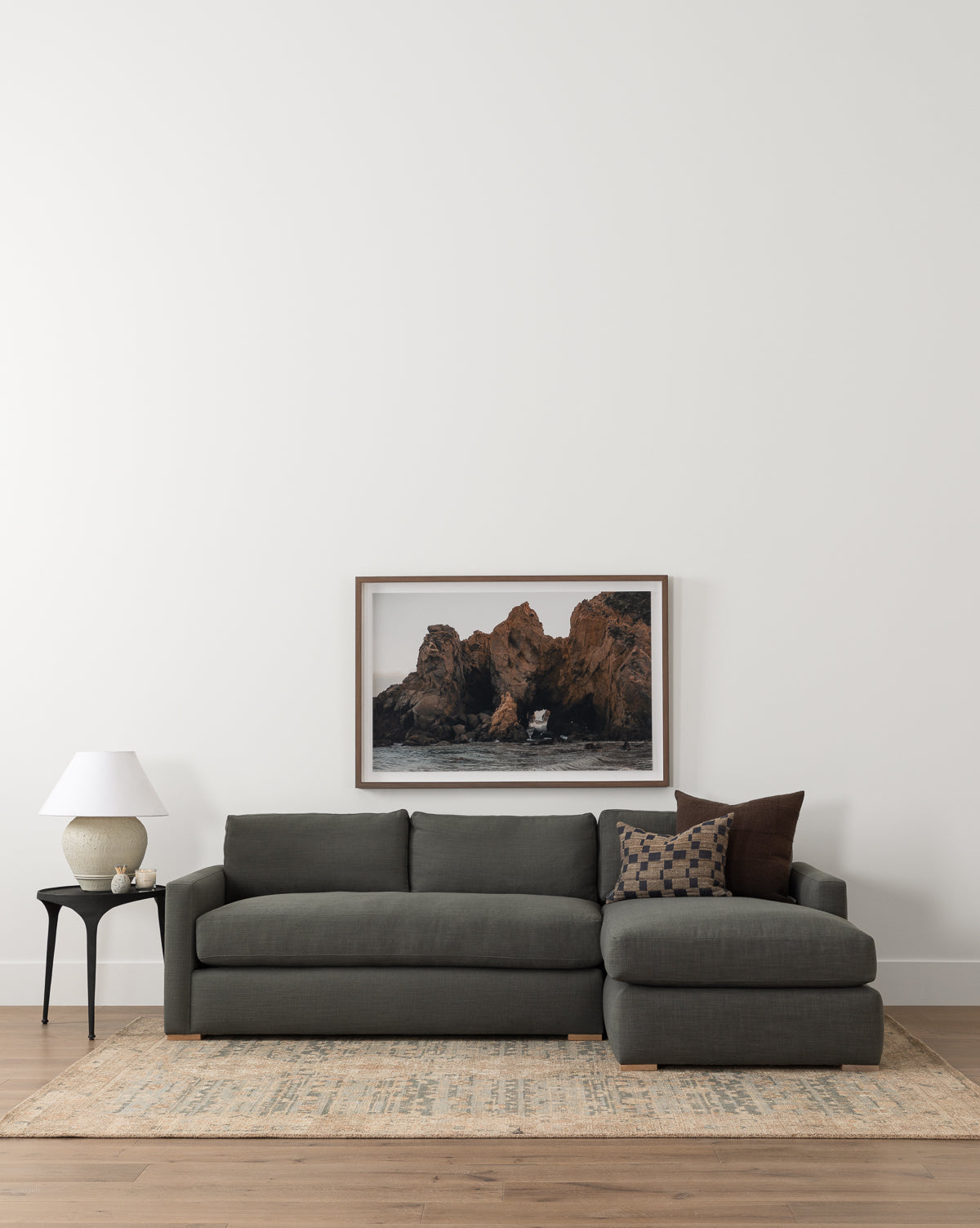 A contemporary living room featuring the Community Macy Upholstered Chaise Sectional 101 in dark gray, patterned throw pillows, a side table with a white lamp and vase, a light rug, and a framed coastal rock photo on a white wall, styled with Vendixs