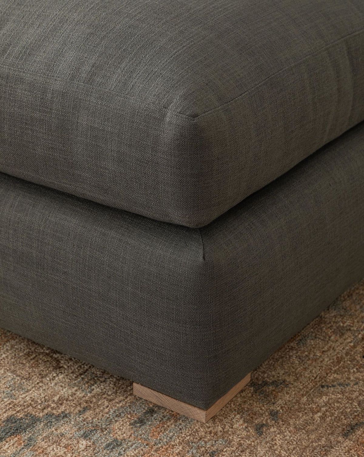 Close-up of the Macy Upholstered Chaise Sectional 101 by Community in dark gray, featuring a plush cushion and low square wooden legs, set on a beige and brown textured rug for a modern look. Vendixs