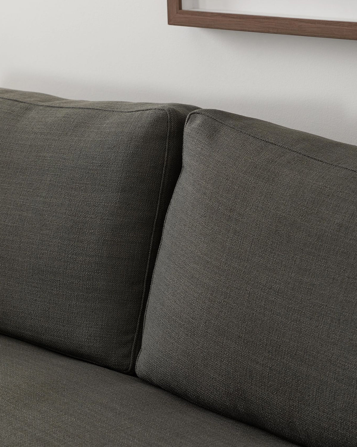 Close-up of the Macy Upholstered Chaise Sectional 101 by Community in dark gray, featuring two seat cushions and a partial picture frame on a white wall in the background. Vendixs