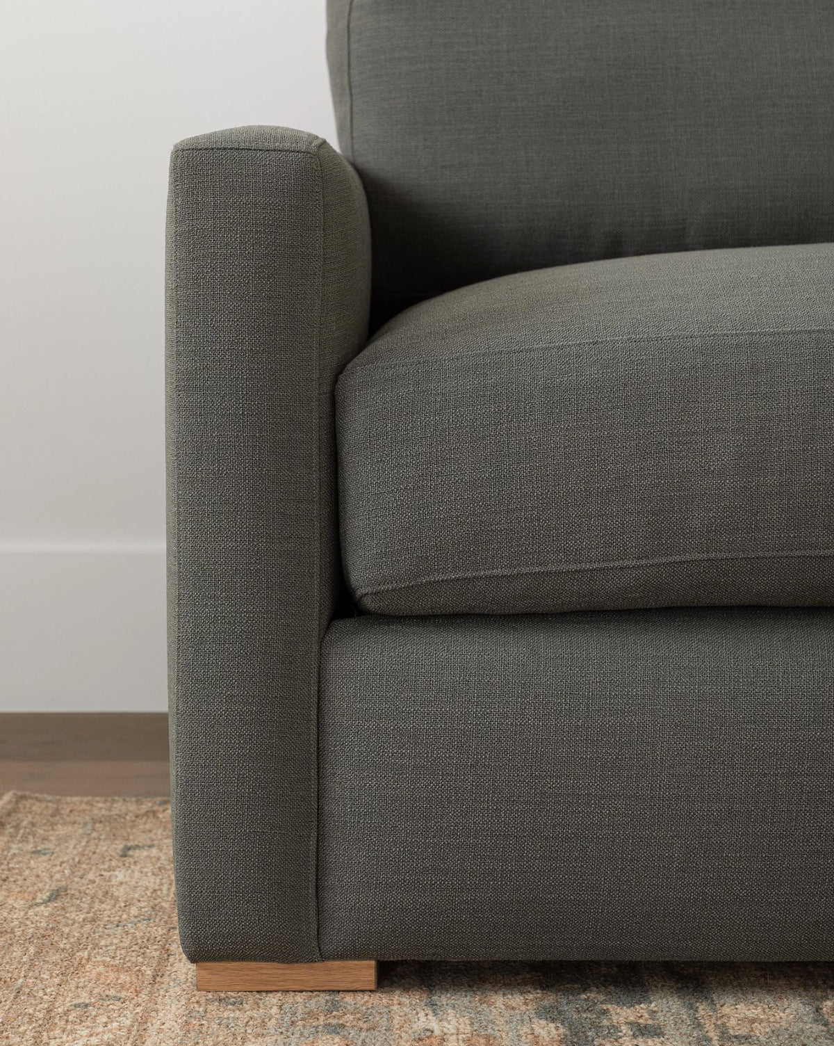 Close-up of the Macy Upholstered Chaise Sectional 101 by Community in dark gray, highlighting its armrest, seat cushion, and wooden leg, with a glimpse of a patterned rug and wooden flooring underneath. Vendixs