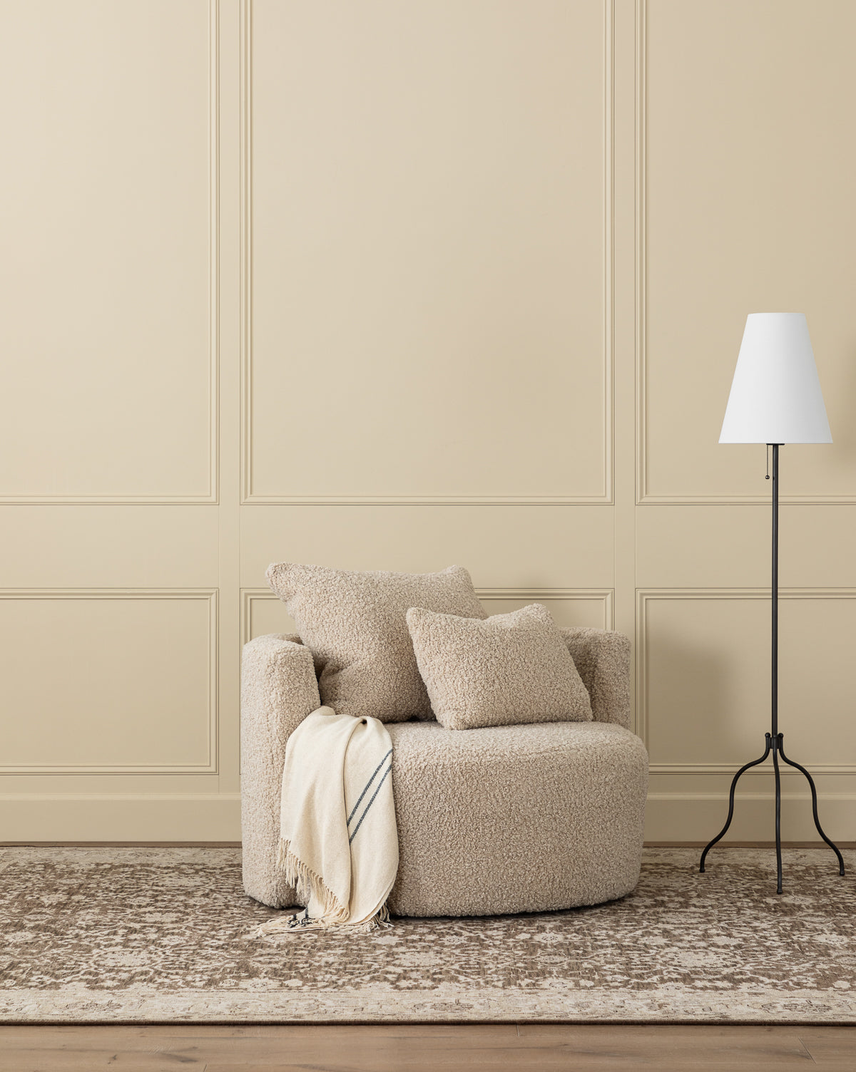 The Banbury Swivel Chair, upholstered in beige and topped with two cushions and a striped throw blanket, sits on a patterned rug beside a white floor lamp against a light beige paneled wall, by McGee & Co.