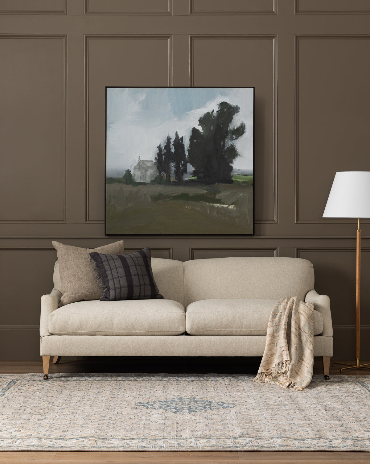 A modern living room showcases the Rowley Sofa 85 by Rowe Fine Furniture, Inc. with customizable fabric, plaid and solid pillows, a plaid throw, floor lamp, gray-blue rug, and landscape painting on a taupe paneled wall—finished with touches from Vendixs